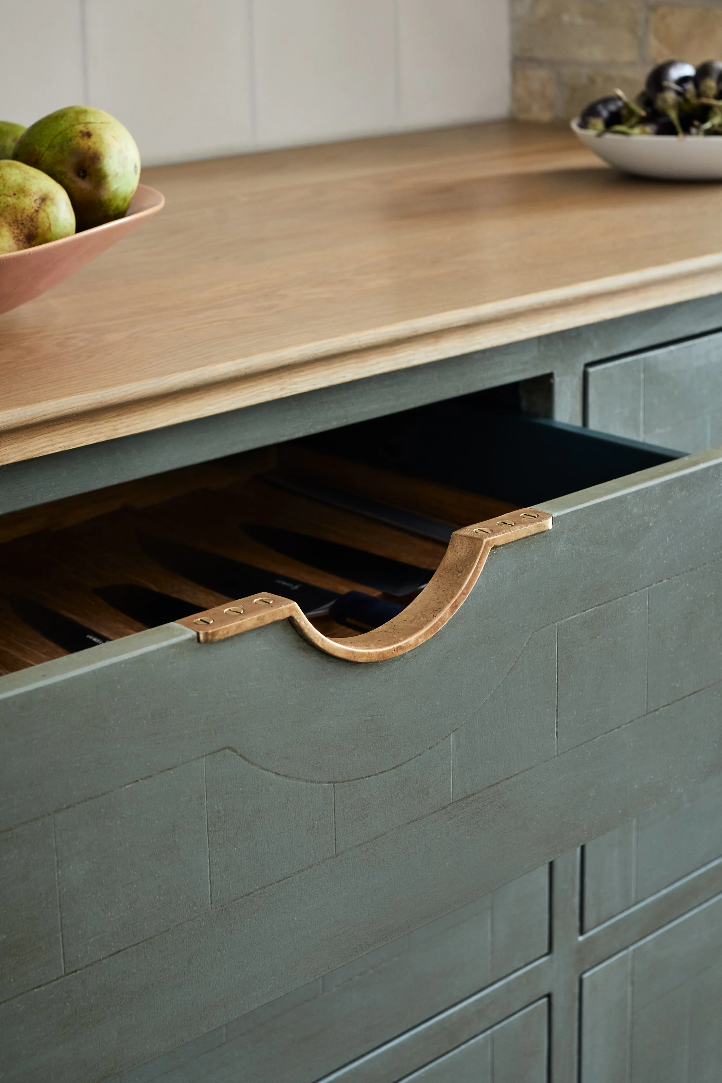 Close-up of a Moray cast bronze pull handle on an open drawer in a green hand-painted kitchen, with knives visible and oak work surface, bowl of pears, and small aubergines in the background.