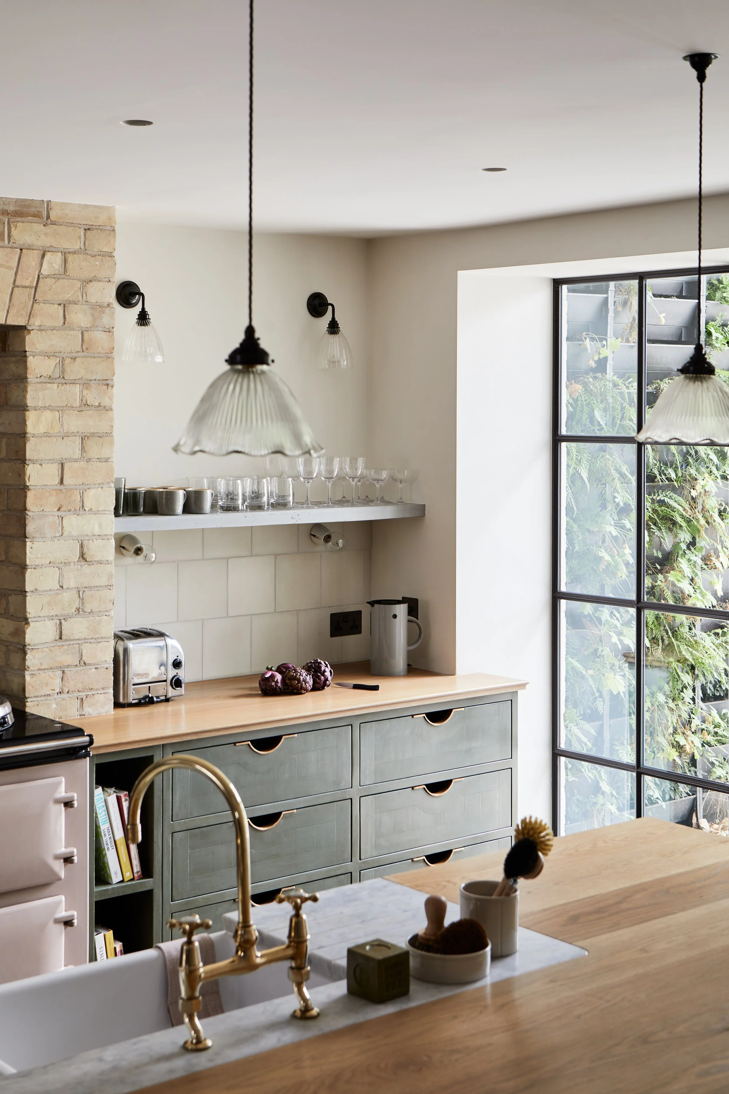 Mid/wide shot of a bank of 6 Moray drawer pulls in a painted green kitchen, with a sink in the foreground, brass mixer tap, pink Aga, and a concrete shelf with glasses in the background.