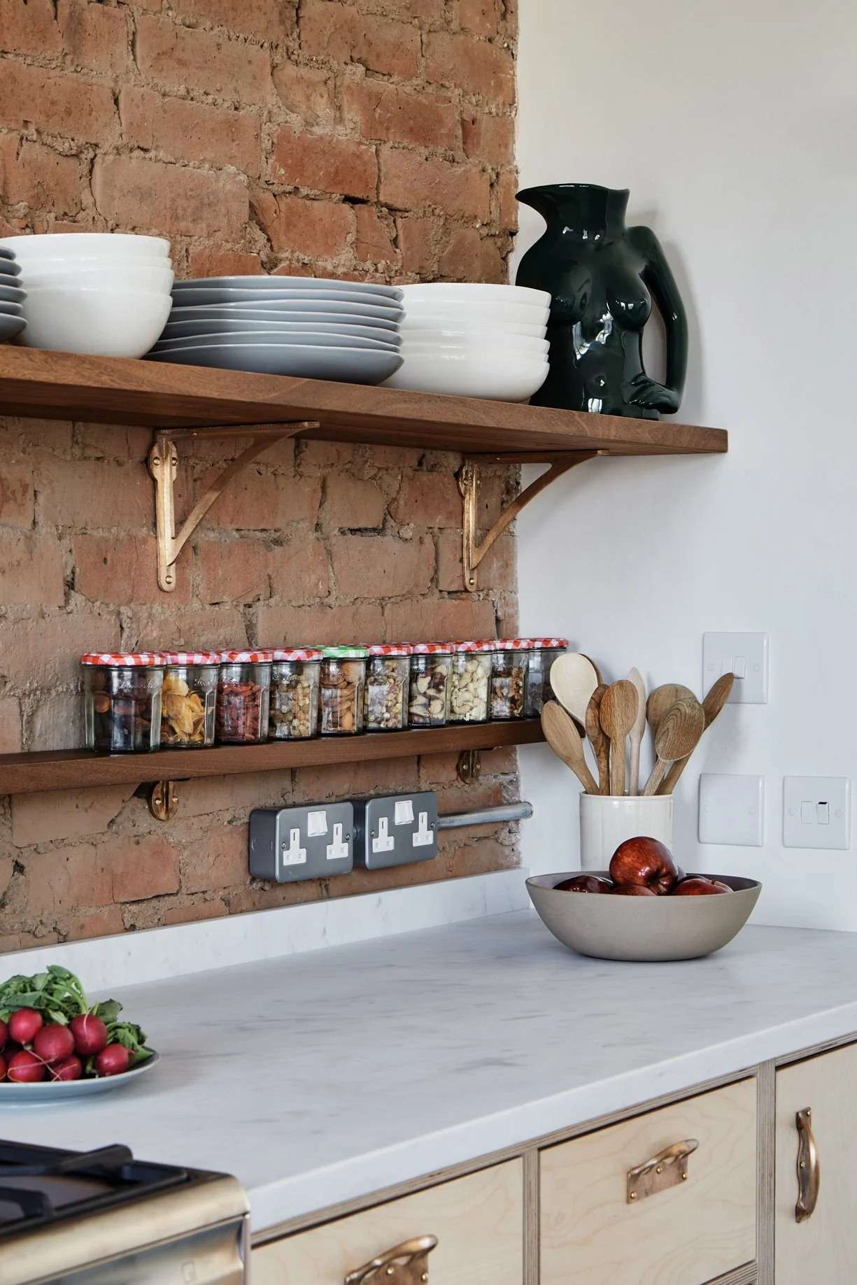 Mid-shot of two 8" Strong Boy Brackets in cast bronze on a brick wall, supporting a timber kitchen shelf with grey plates. Below, a smaller 4" shelf holds jars of nuts.