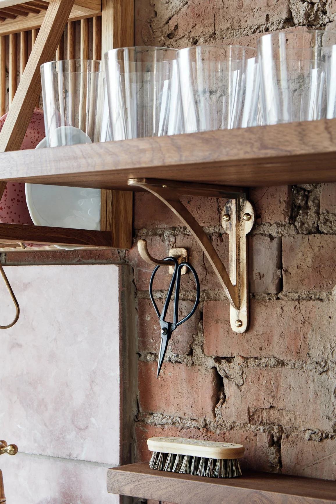 Close-up of an 8" Strong Boy Bracket in cast bronze on a brick wall, supporting a timber kitchen shelf. Drinking glasses sit on the shelf, with scissors hanging from a hook below.
