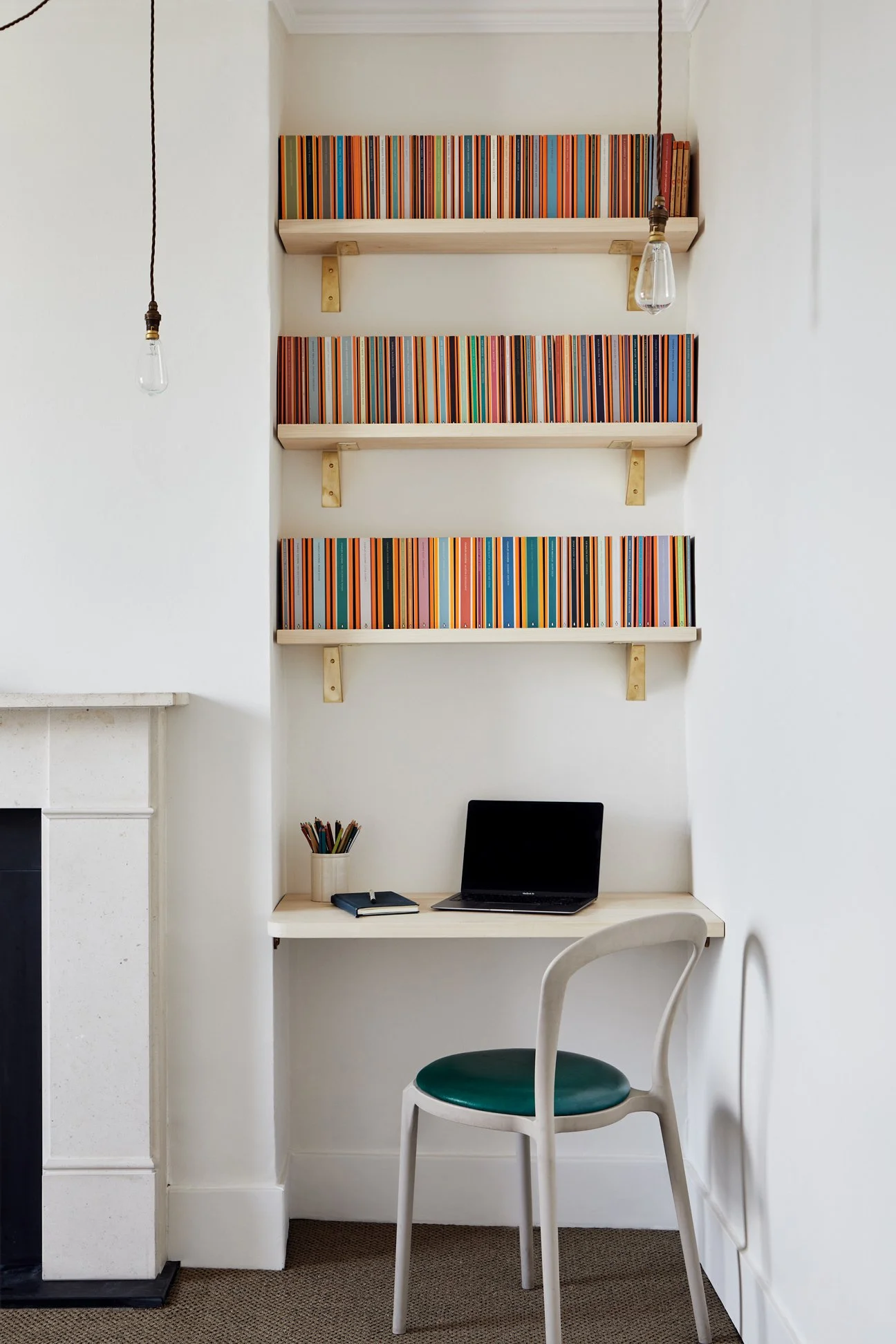 Straight-on view of a workspace with a timber desk and a green leather-seated chair below three shelves supported by brass folded brackets. The edge of a fireplace is visible to the left.