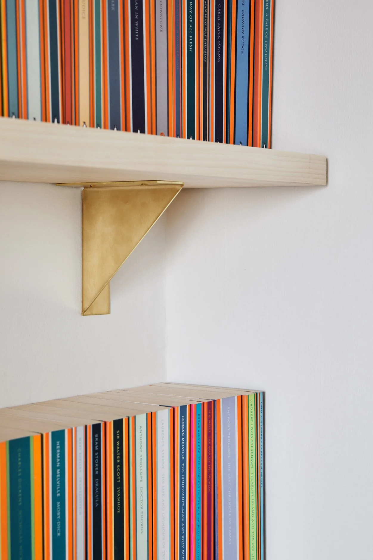 Close-up of the left-hand brass folded bracket supporting a timber shelf filled with Penguin Classics books. The unlacquered brass develops a rich patina over time, adding warmth to the modern design.