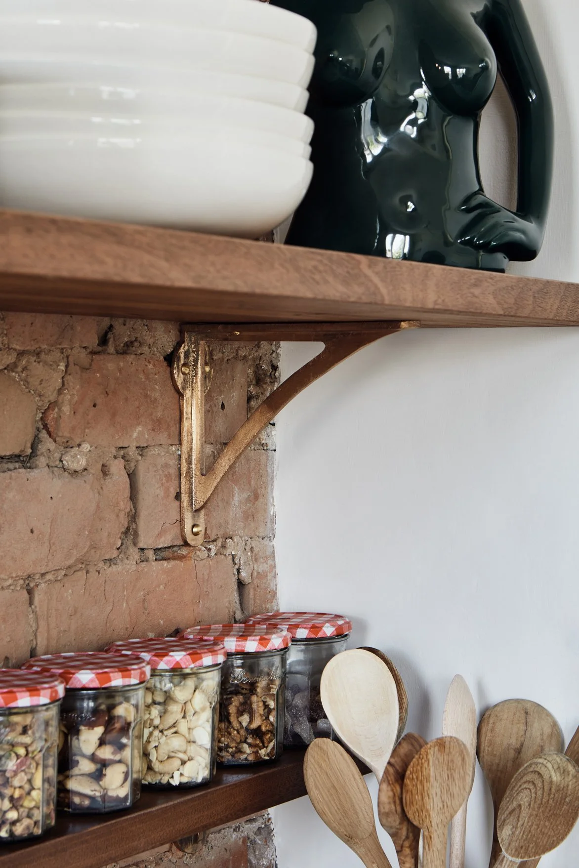 Close-up of an 8" Strong Boy Bracket in cast bronze on a brick wall, supporting a timber kitchen shelf. Below, a smaller 4" shelf holds jars of nuts.