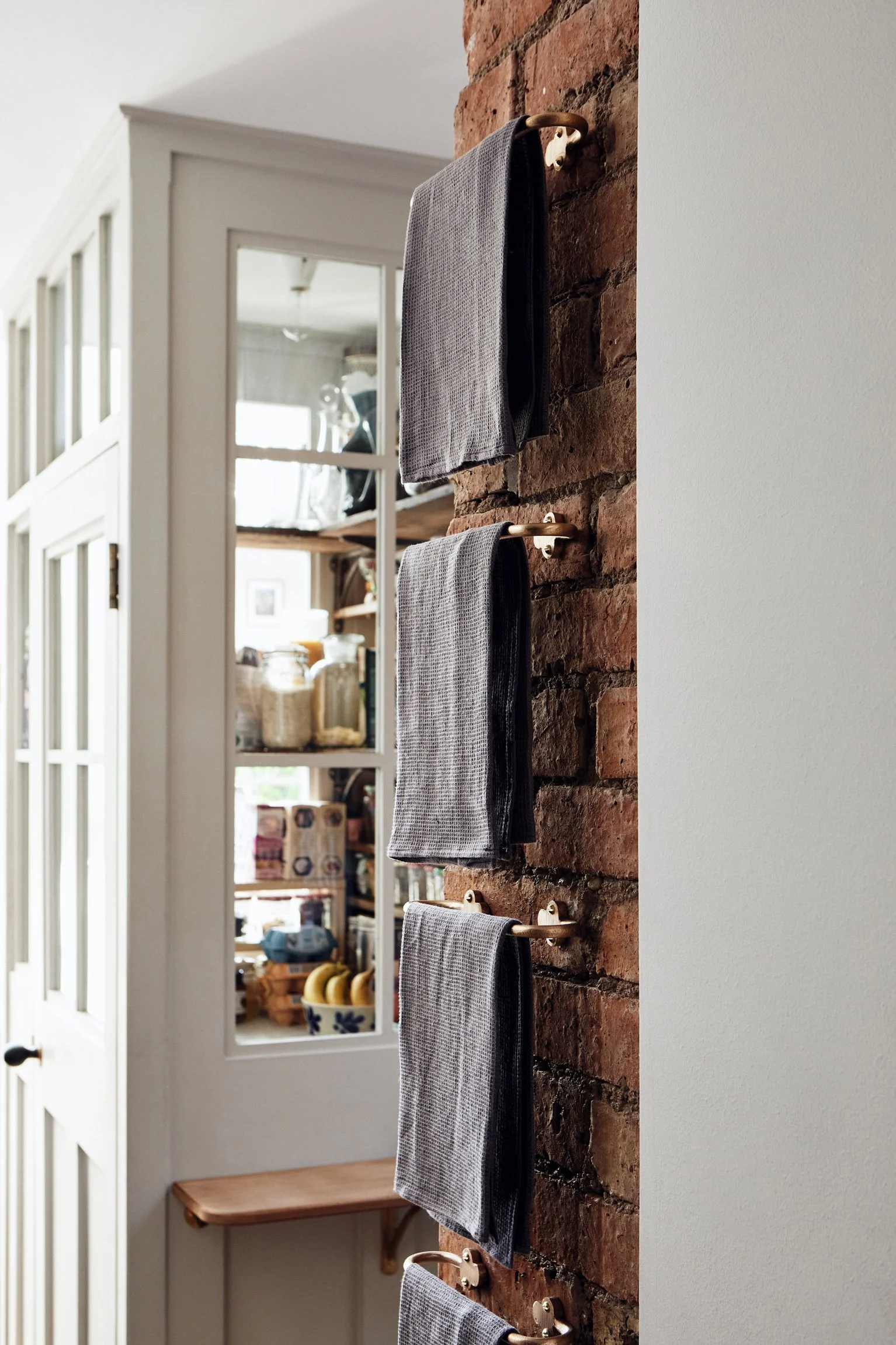 Hand Towel Rail, Cast Bronze side view of 3 rails fixed to a brick wall, hanging one above the other with waffle towels. Semi-glazed larder in the background. Made in raw bronze.