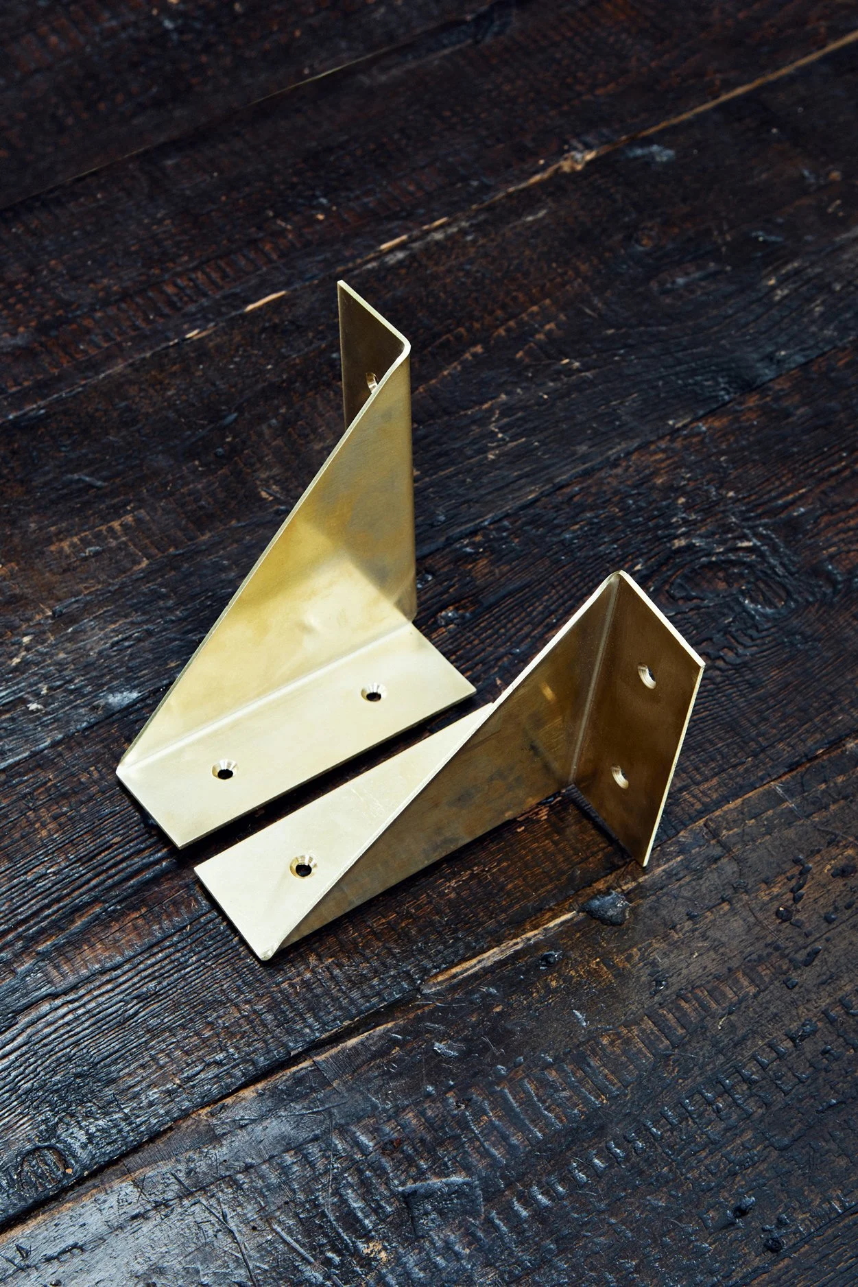A pair of brass folded brackets resting on a timber display board, positioned side by side. The unlacquered brass finish will develop a rich patina over time.