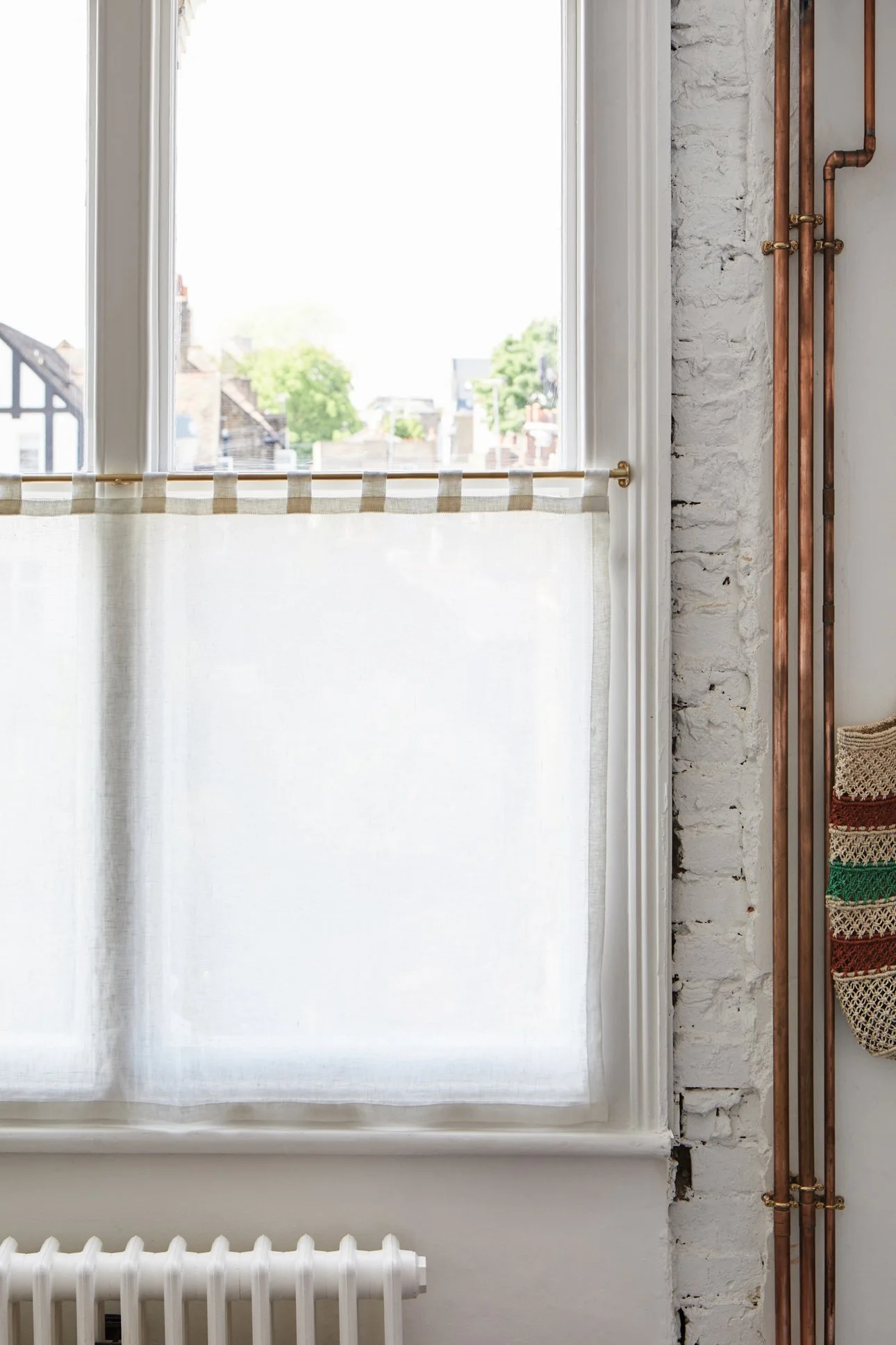 Unlacquered Brass Café Curtain Rail Set mid shot of right side of triple box sash window, curtain covers bottom sash, exposed copper pipe run vertically to the right.