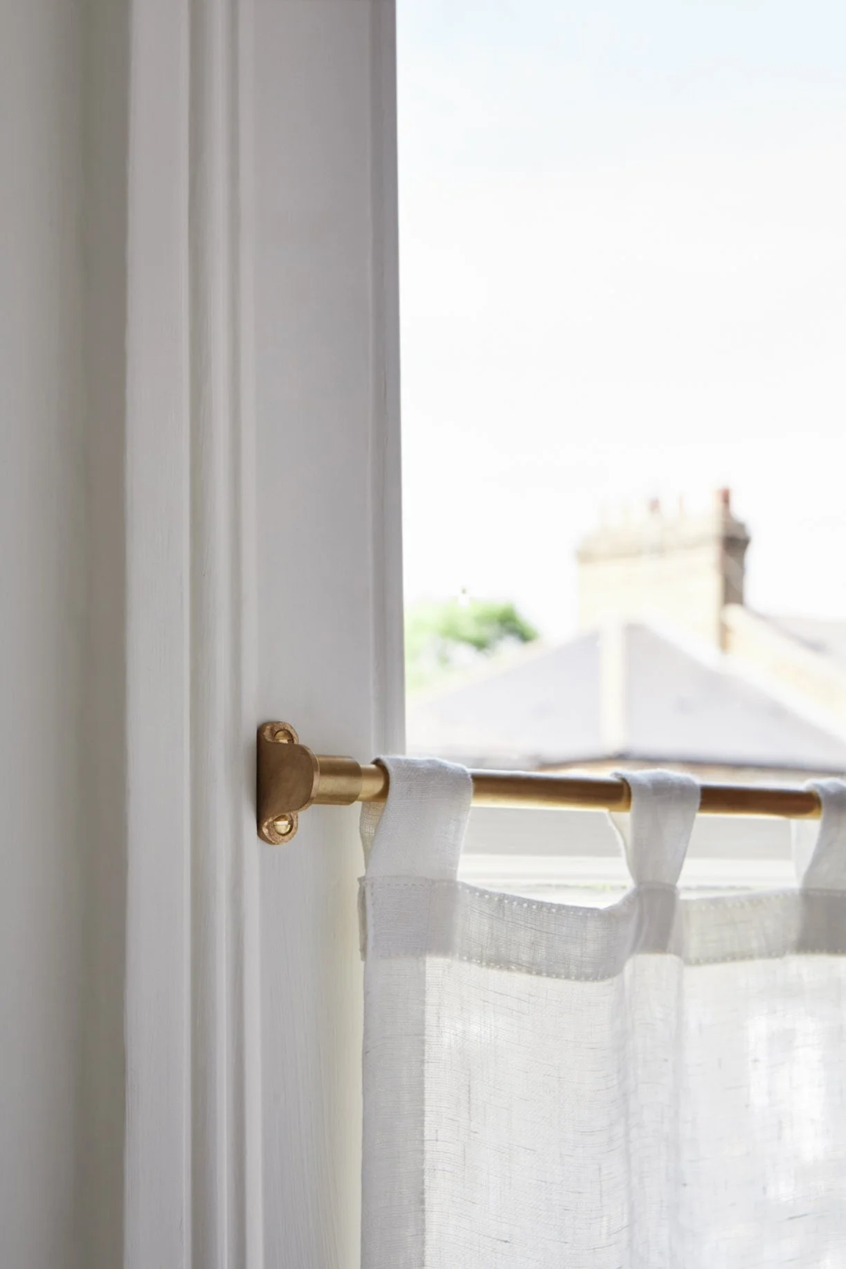 Café Curtain End Brackets - Closer, side-on shot of the bracket fixed to the window frame, with a linen café curtain hanging, and chimneys out of focus in the distance.