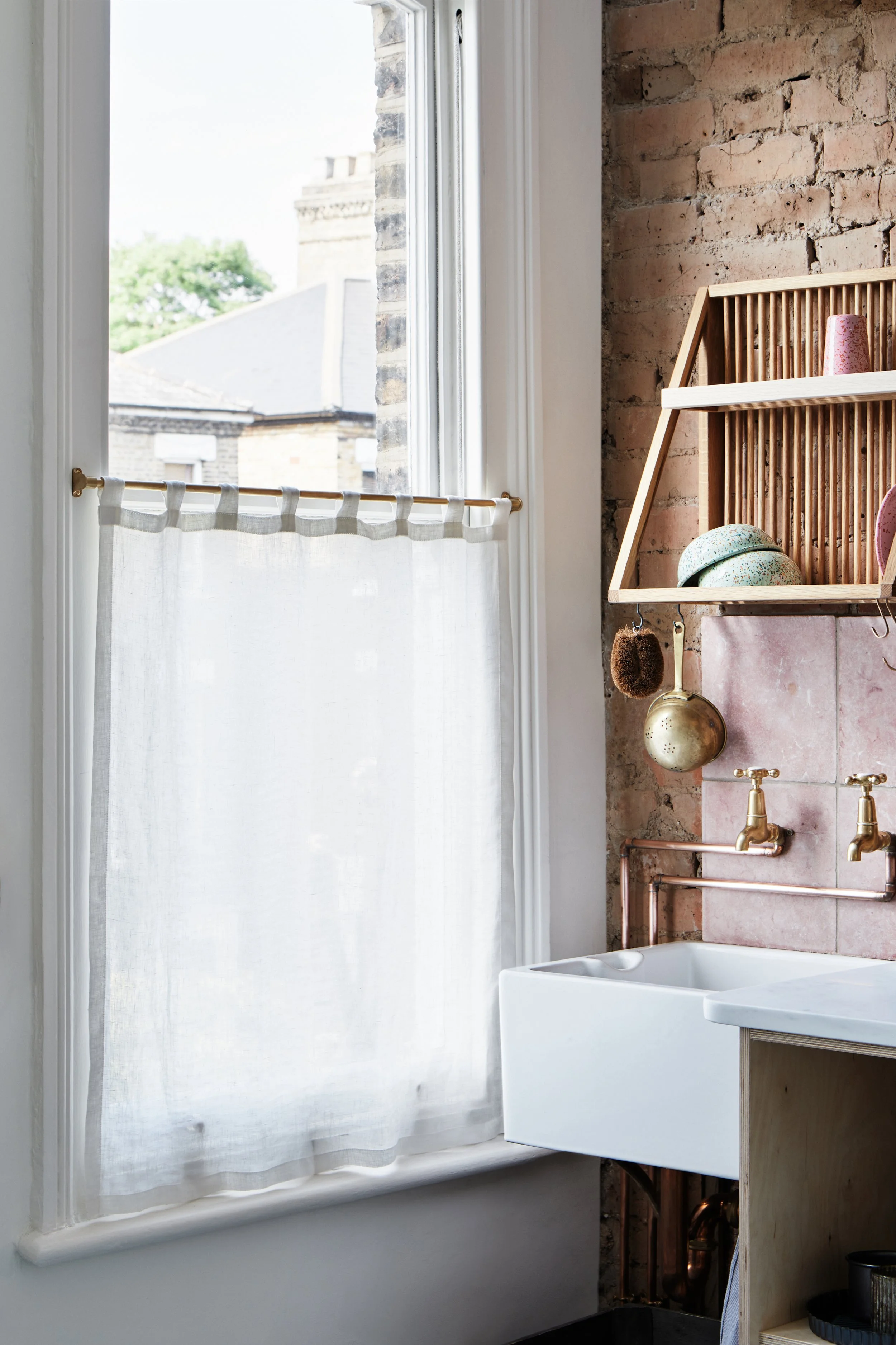 Brass Café Curtain Rail Set Mid shot of a single sash window with Linen curtain hung halfway, showing the brass rail, Belfast sink, wooden draining rack, brass taps, and pink splashback tiles