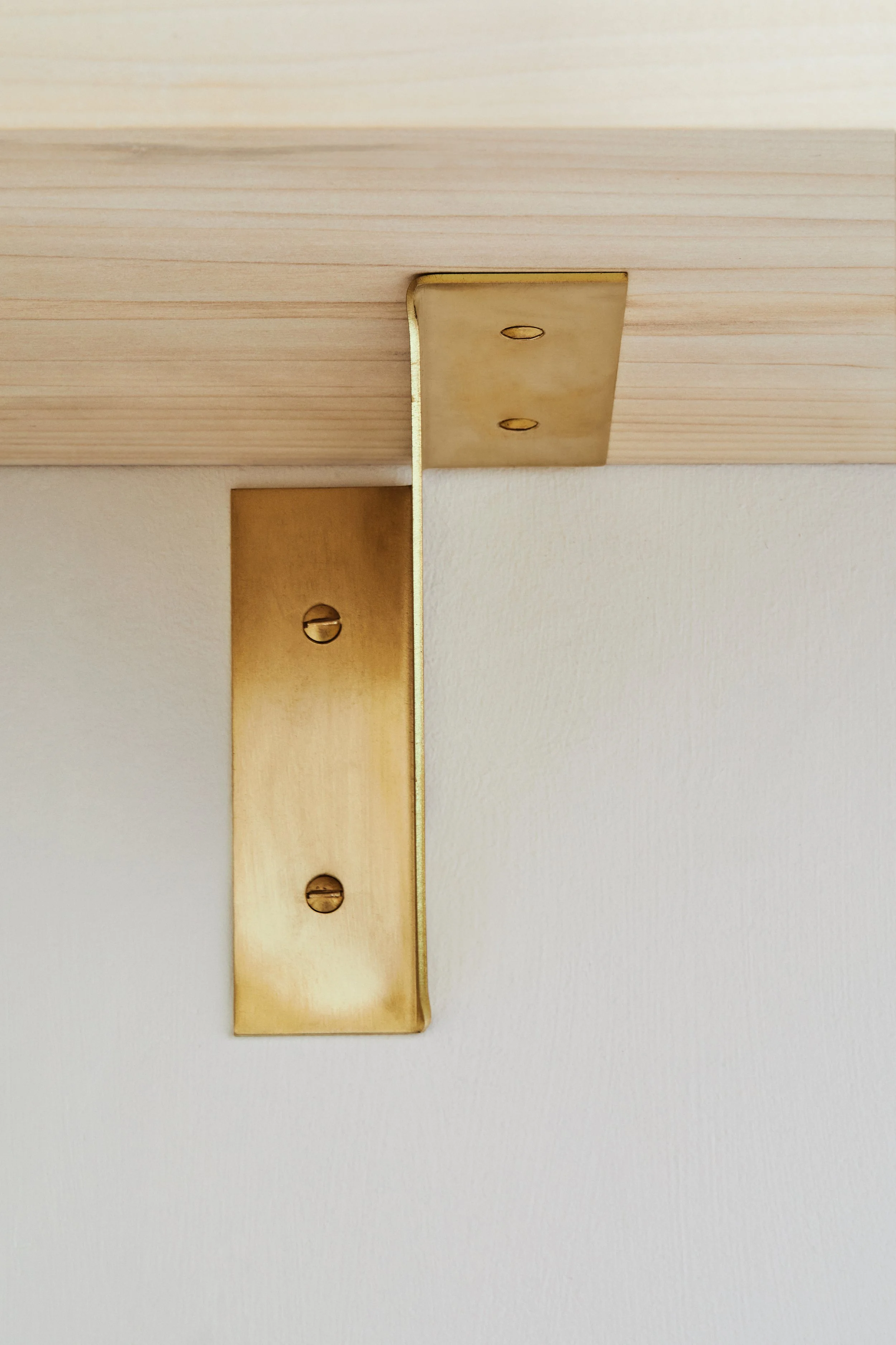 Close-up of the right-hand brass folded bracket with brass fixings on a pale timber shelf against a linen wash wall. The unlacquered brass develops a rich patina over time.