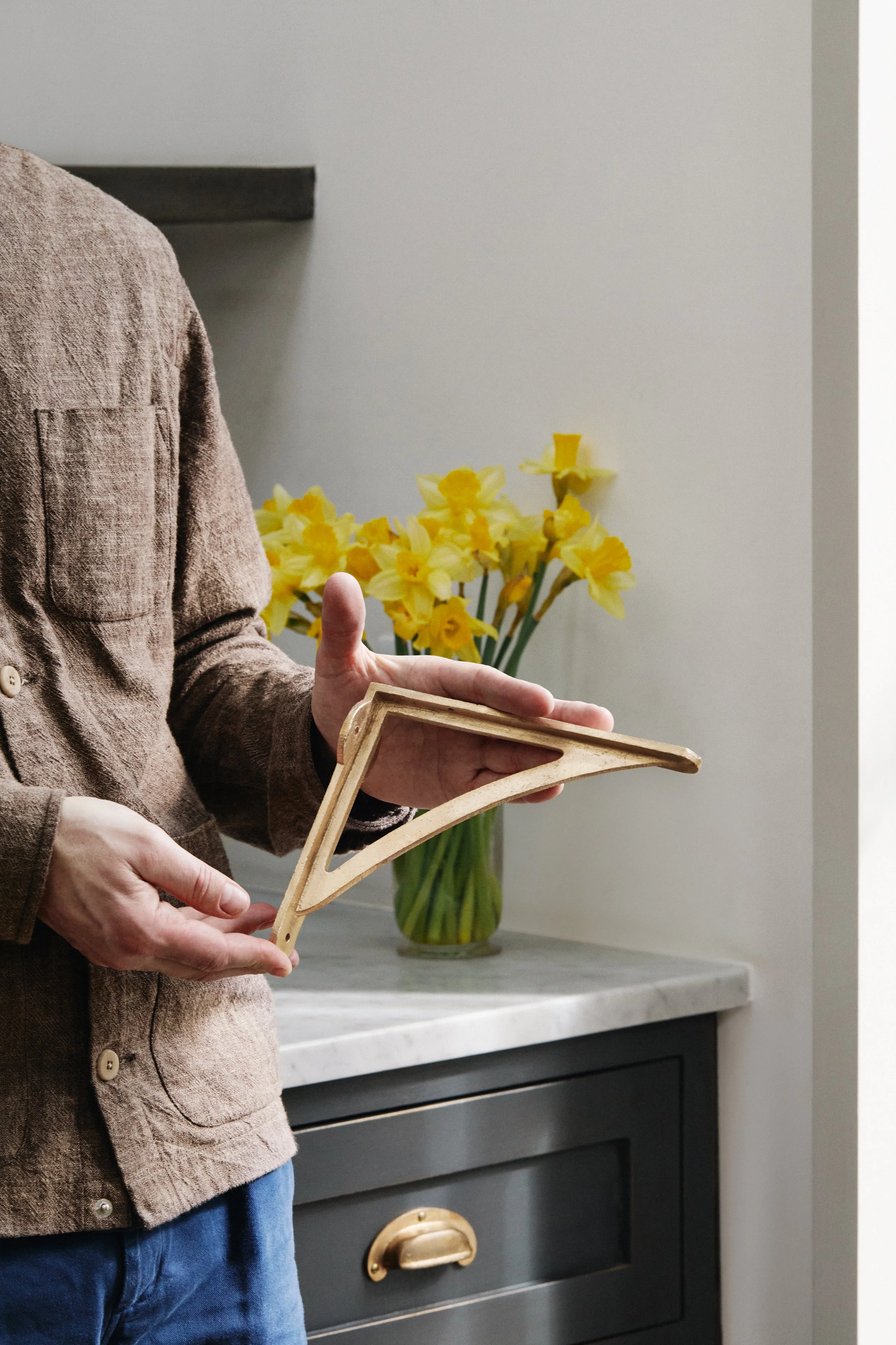 Mark holding an 8" Strong Boy Bracket in cast bronze in his kitchen. A marble surface and daffodils are visible in the background