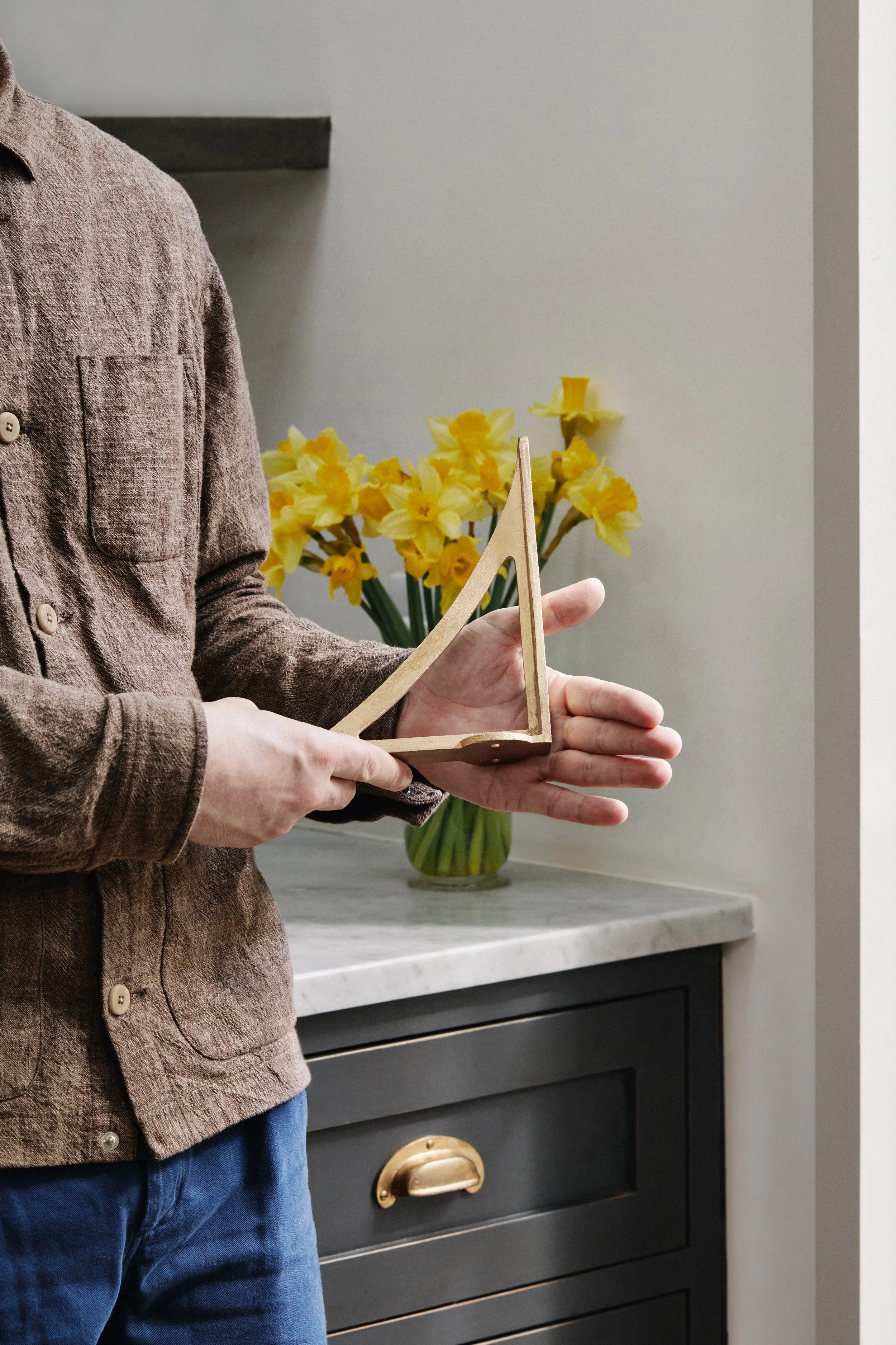 Mark holding an 8" Strong Boy Bracket in cast bronze at a different angle in his kitchen. A marble surface and daffodils are visible in the background