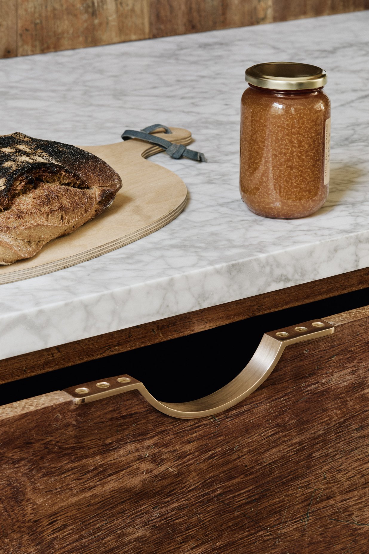 Close-up of a Moray drawer pull in a reclaimed iroko kitchen, with a marble work surface, a jar of honey, a breadboard, and a loaf of sourdough in the background.