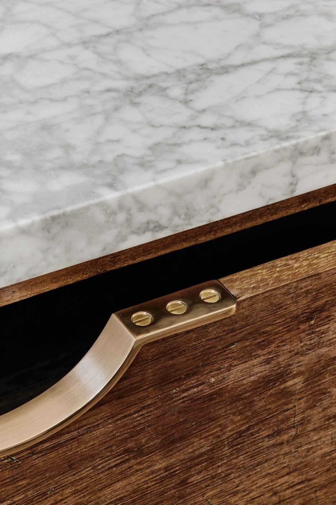 Very close-up of 3 screws fitting a Moray drawer pull, with the drawer front ajar in a reclaimed iroko kitchen, and a grey marble kitchen work surface visible at the top.