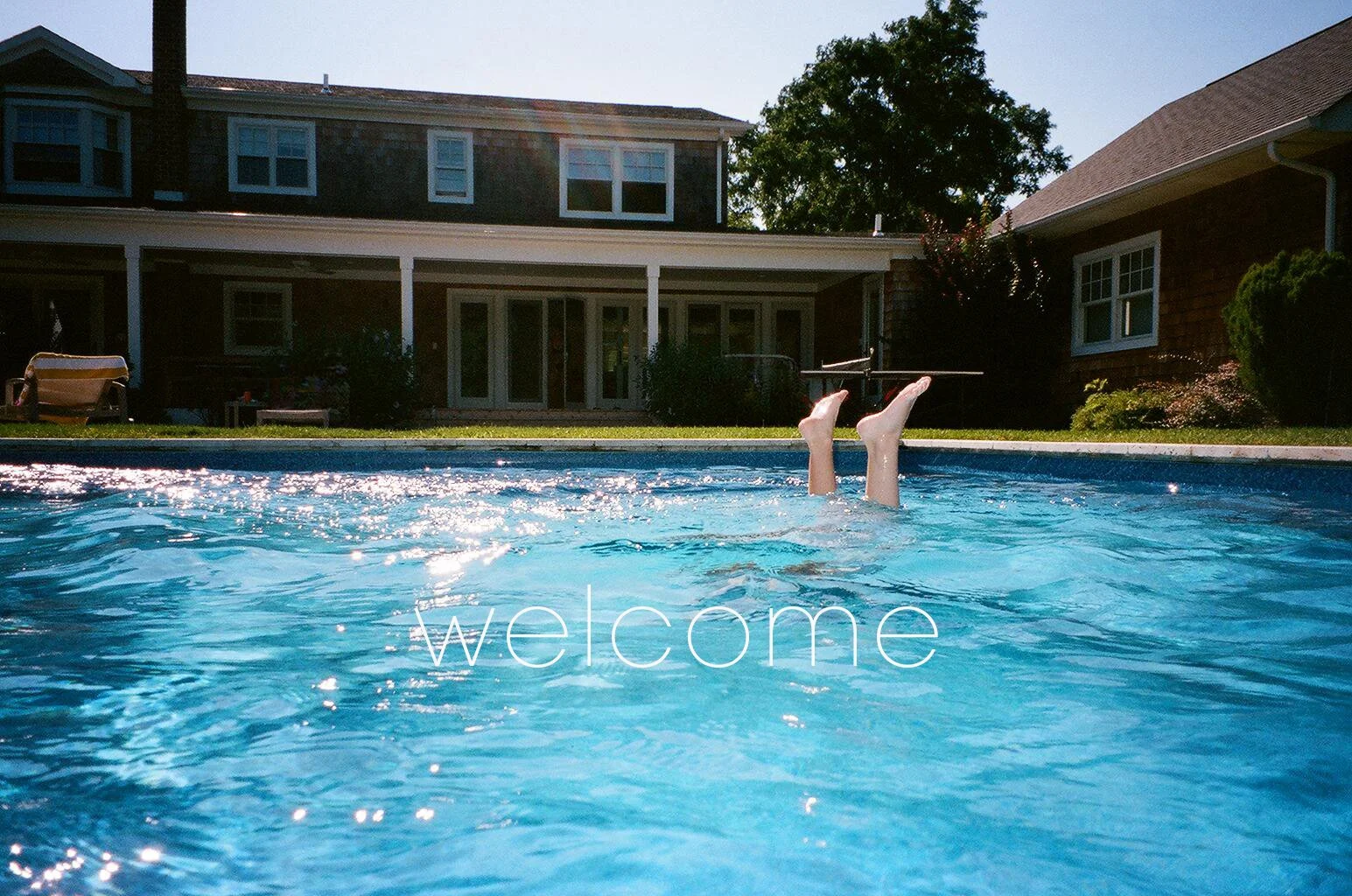 a shimmering blue pool is in the foreground. a child, doing an underwater handstand, her feet just visible above the water. a gracious and expansive house is in the background.