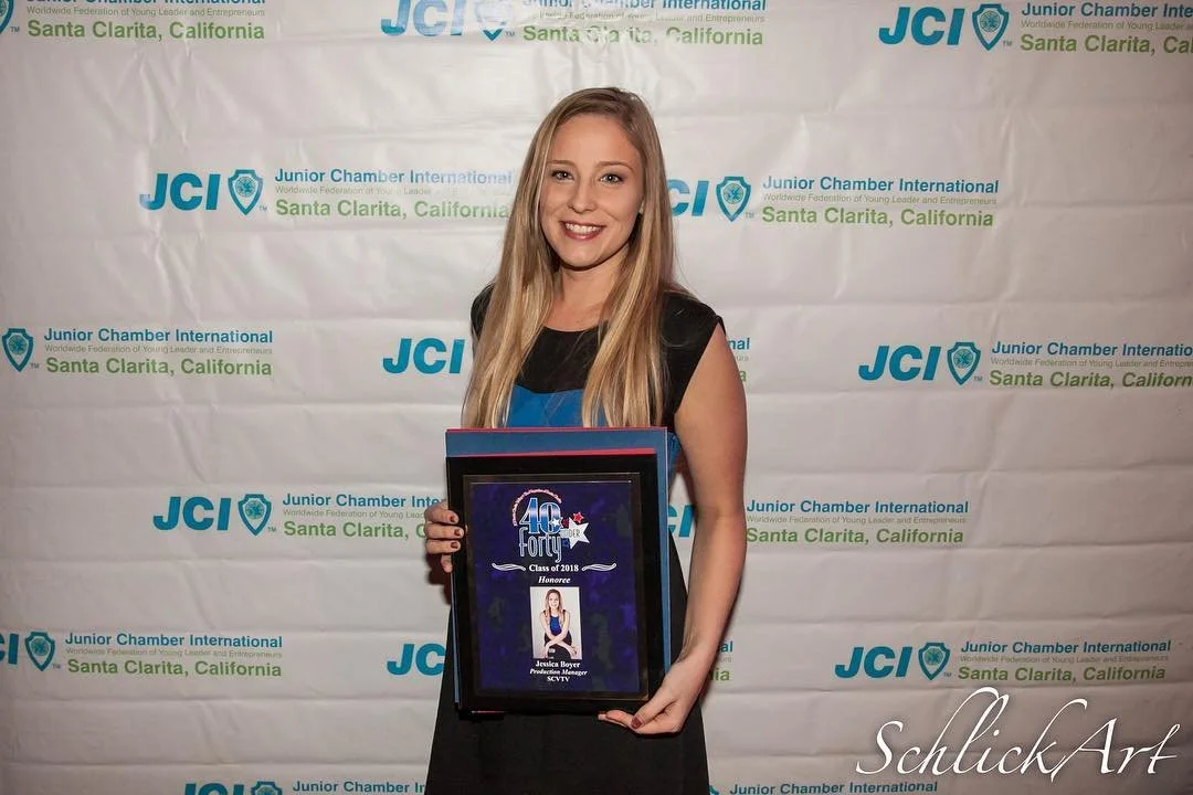 Young woman in black dress holding a framed award plaque in front of a white step and repeat banner with blue and green logos for Junior Chamber International Santa Clarita, California, smiling at the camera.
