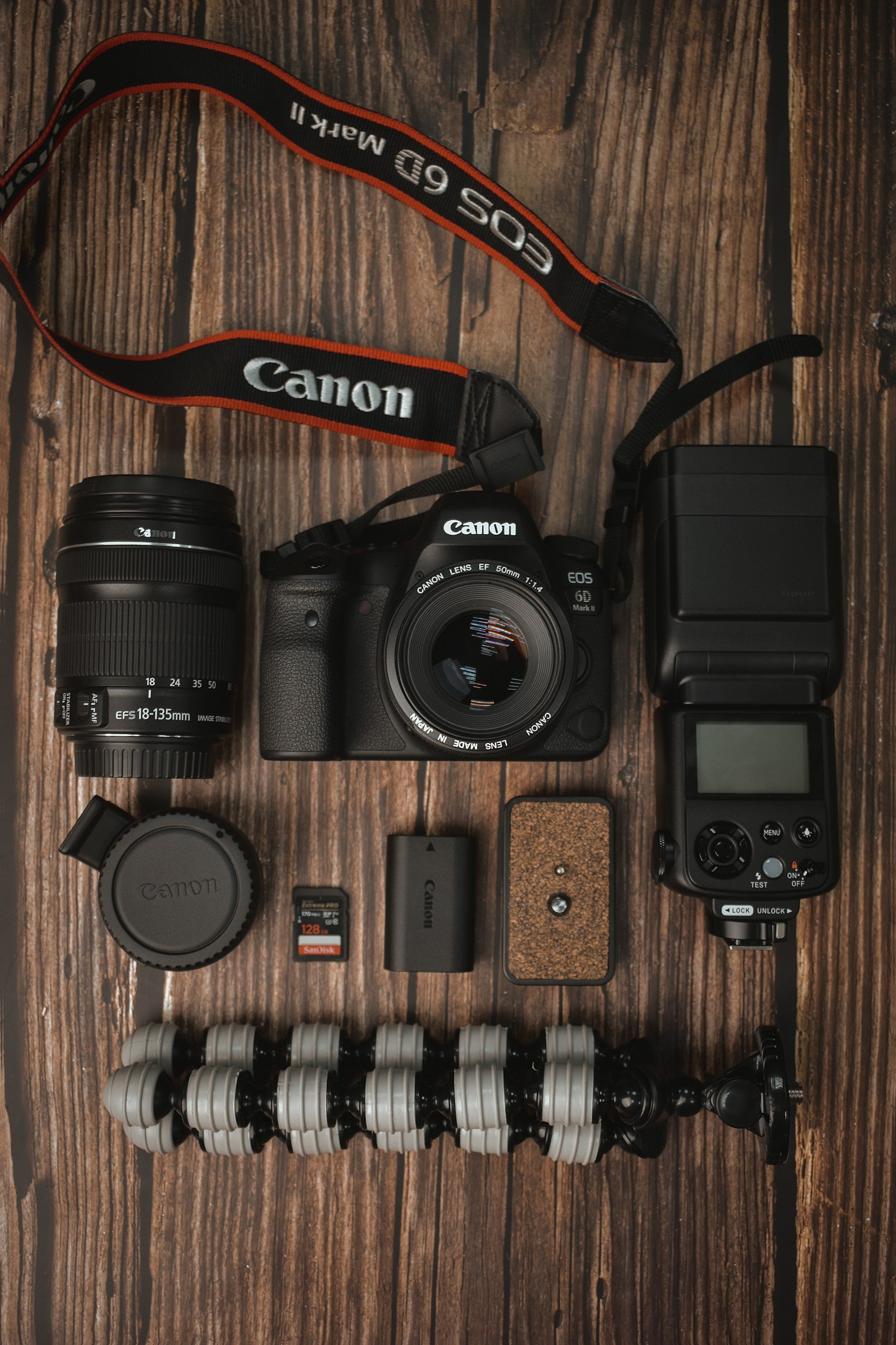 Photo of a Canon camera kit on a wooden surface. It includes a Canon EOS 6D Mark II camera body with a lens attached, an additional Canon zoom lens, a camera strap, a Canon lens cap, an SD memory card, a camera battery, a battery charger, a small wooden object, and a flexible tripod.