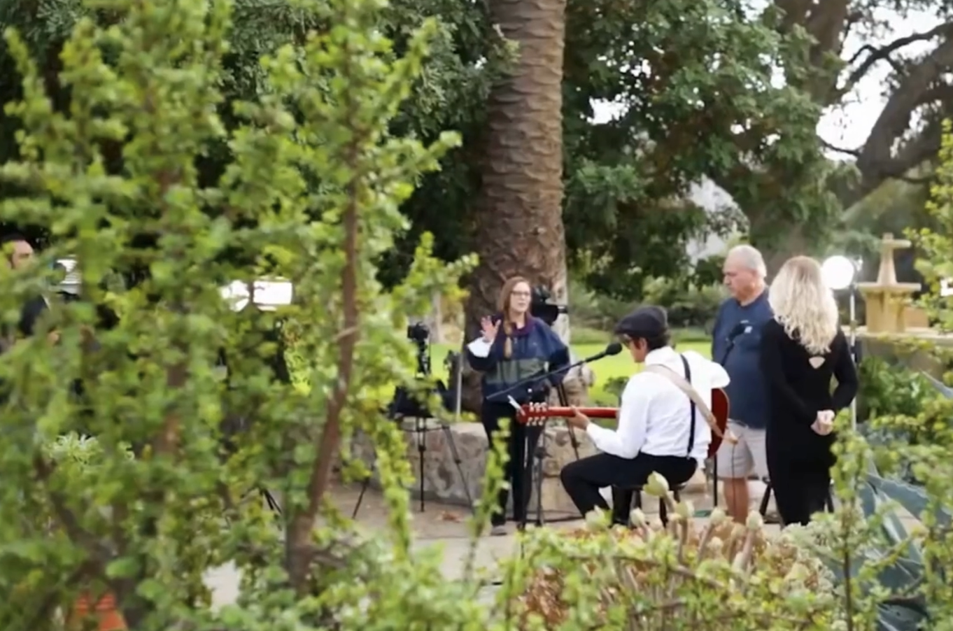 People performing live music outdoors with a singer, guitarist, and onlookers in a garden with large trees and greenery.