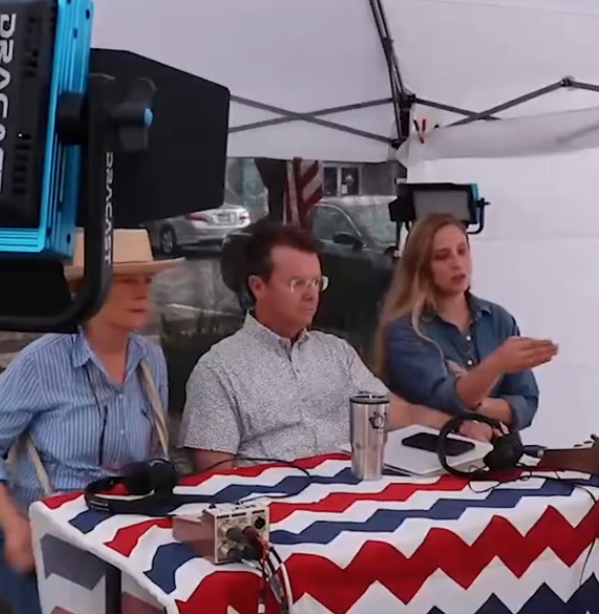 Three people sitting at a table with a red, white, and blue tablecloth, inside a white tent with camera equipment around.