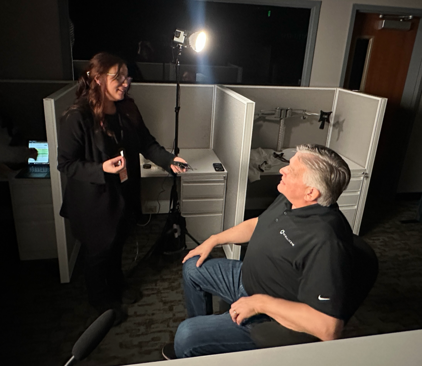 A woman standing and smiling while talking to an older man sitting in a chair, in a dimly lit room with partitions and tech equipment, possibly during an interview or recording session.