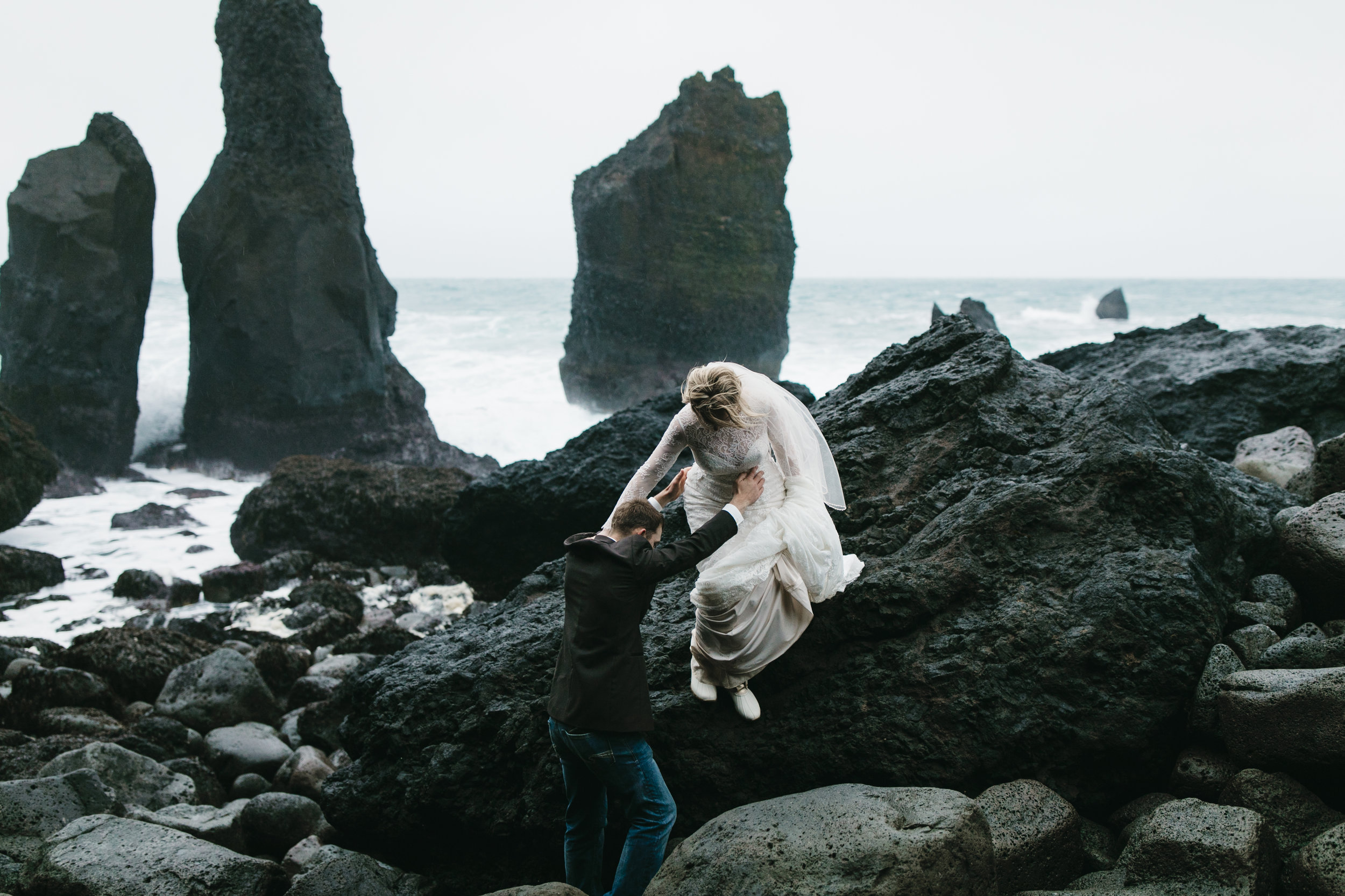 A groom helps his adventurous bride down from rocks on Reykjanes peninsula while Iceland Elopement photographers Colby and Jess capture their adventure.