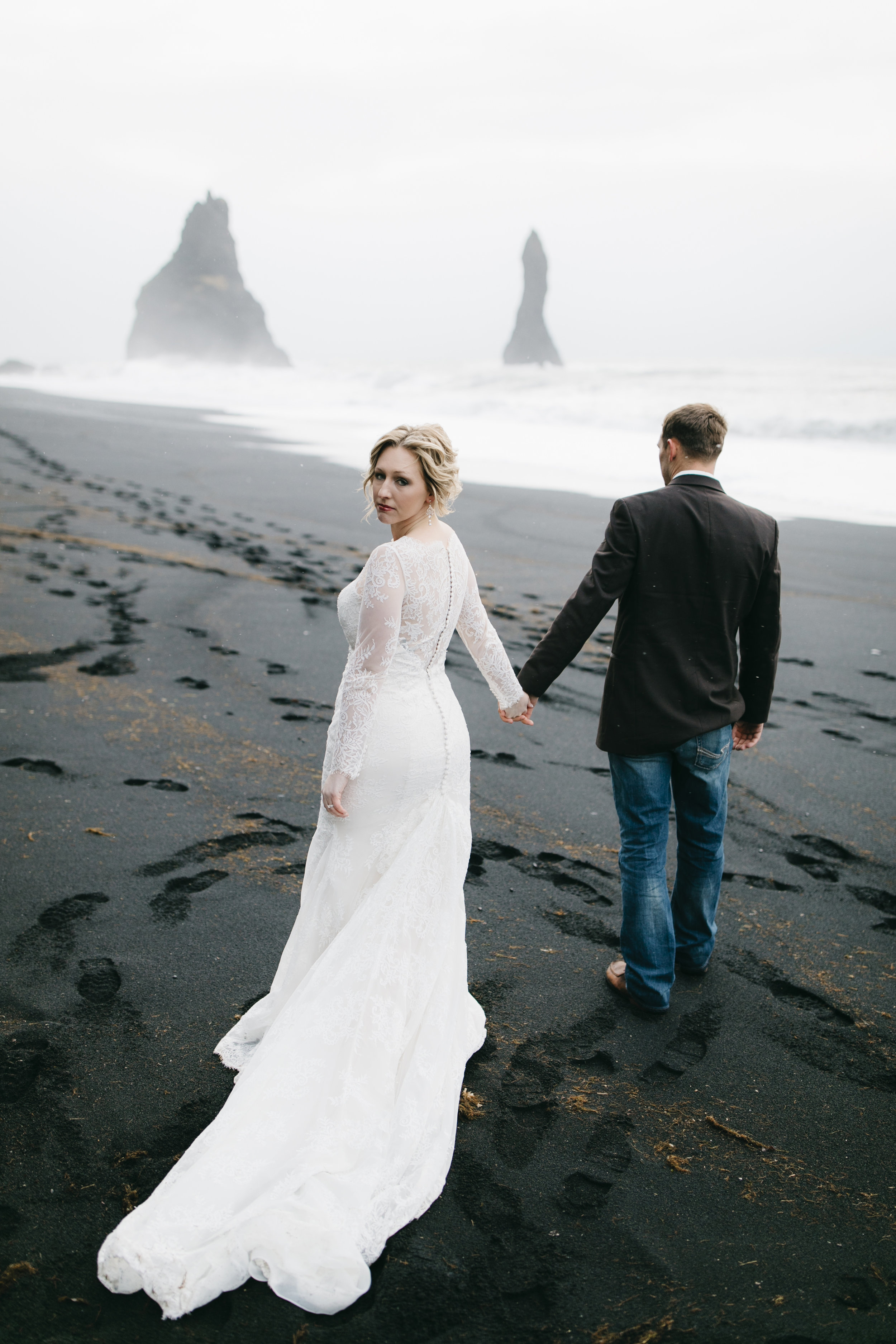 A beautiful bride walks with her groom on a black sand beach in Vik, looking over her shoulder at Iceland Wedding Photographers Colby and Jess, colbyandjess.com