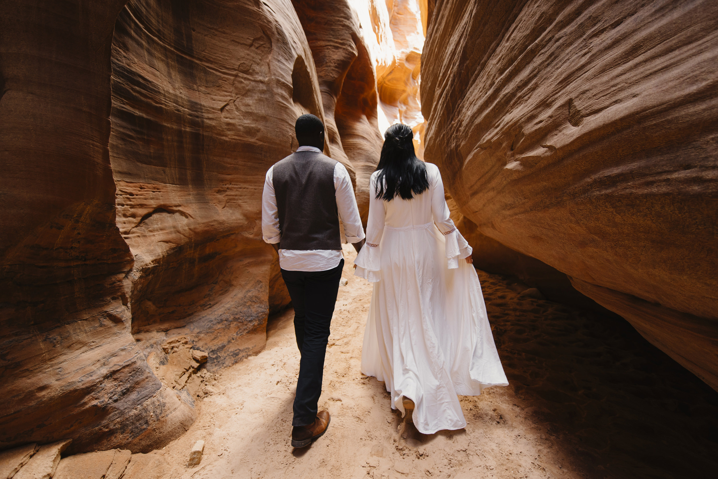 A couple walks holding hands through a slot canyon during their Antelope Canyon Adventure Engagement Photography Session by Utah Destination Elopement Photographer Colby and Jess