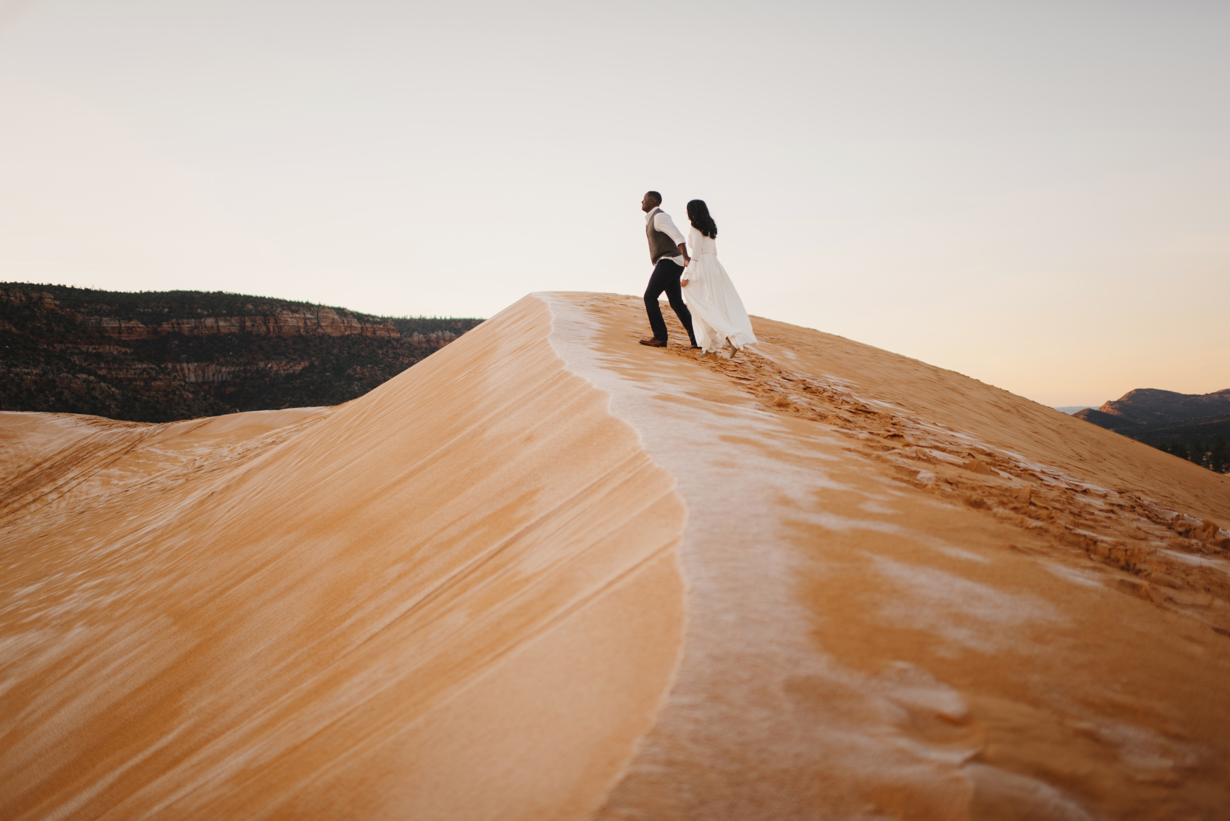 A couple climbs a sand dune during their Coral Pink Sand Dunes Engagement Photography Session with Utah Destination Elopement Photographer Colby and Jess