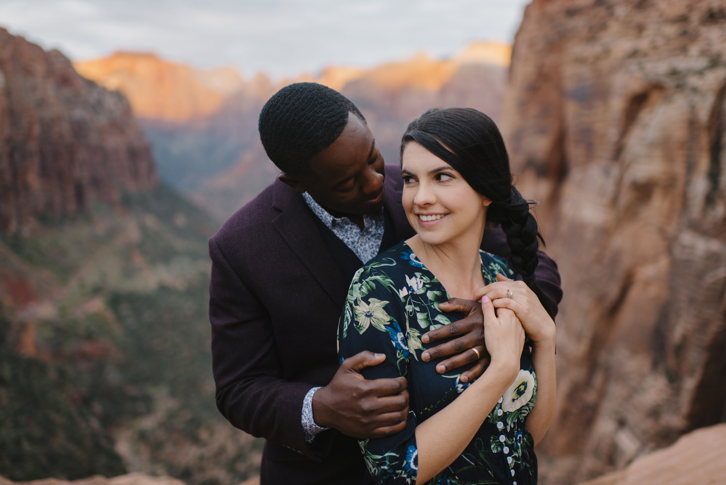 A woman is held by her husband during their Angels Landing Engagement Photography Session with Zion National Park Adventure Elopement Photographer Colby and Jess