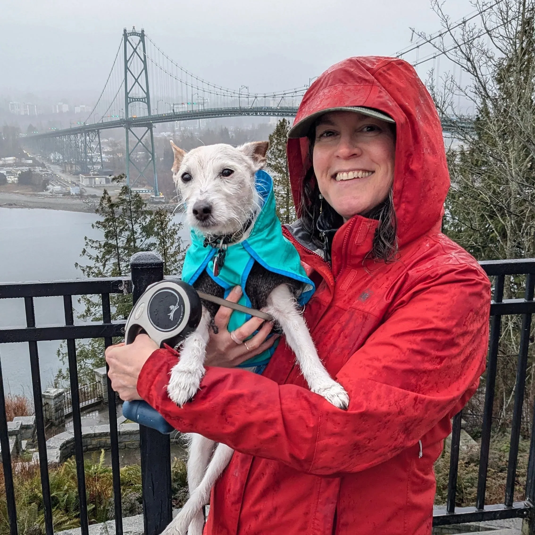Nicole in an orange raincoat holding her little white dog, Dodge, who is in a blue raincoat