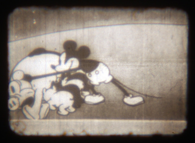 A close-up of the film shows Mickey Mouse picking up a pig while piglets try to get some milk from their mother.