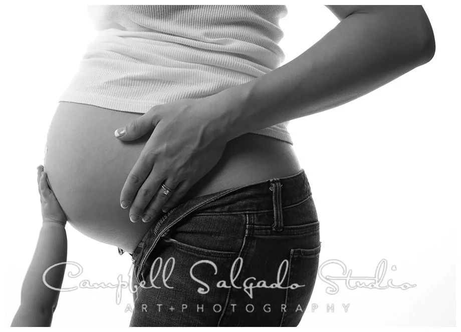B&W portrait of baby bump with siblings hand on white background at Campbell Salgado Studio.
