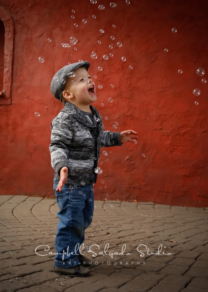 Portrait of child playing with bubbles on red stucco background by child photographers at Campbell Salgado Studio, Portland, Oregon.