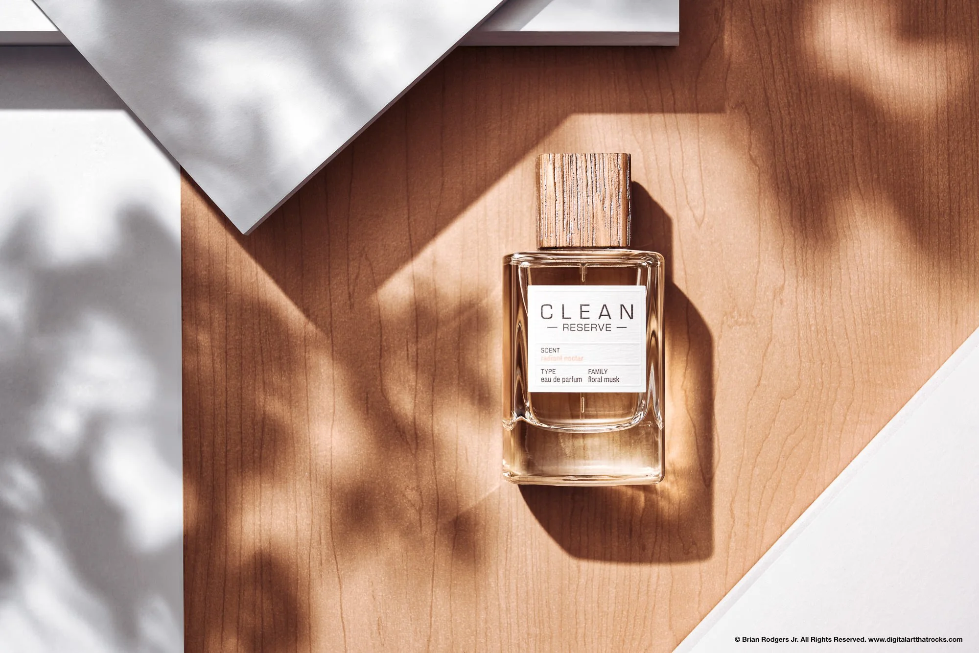 Professional product photography hero shot of a fragrance bottle with a wooden cap, sitting on a warm wooden surface and framed by angular shadows and white props, highlighting the natural, sunny aesthetic. High End product photographer for fragrance