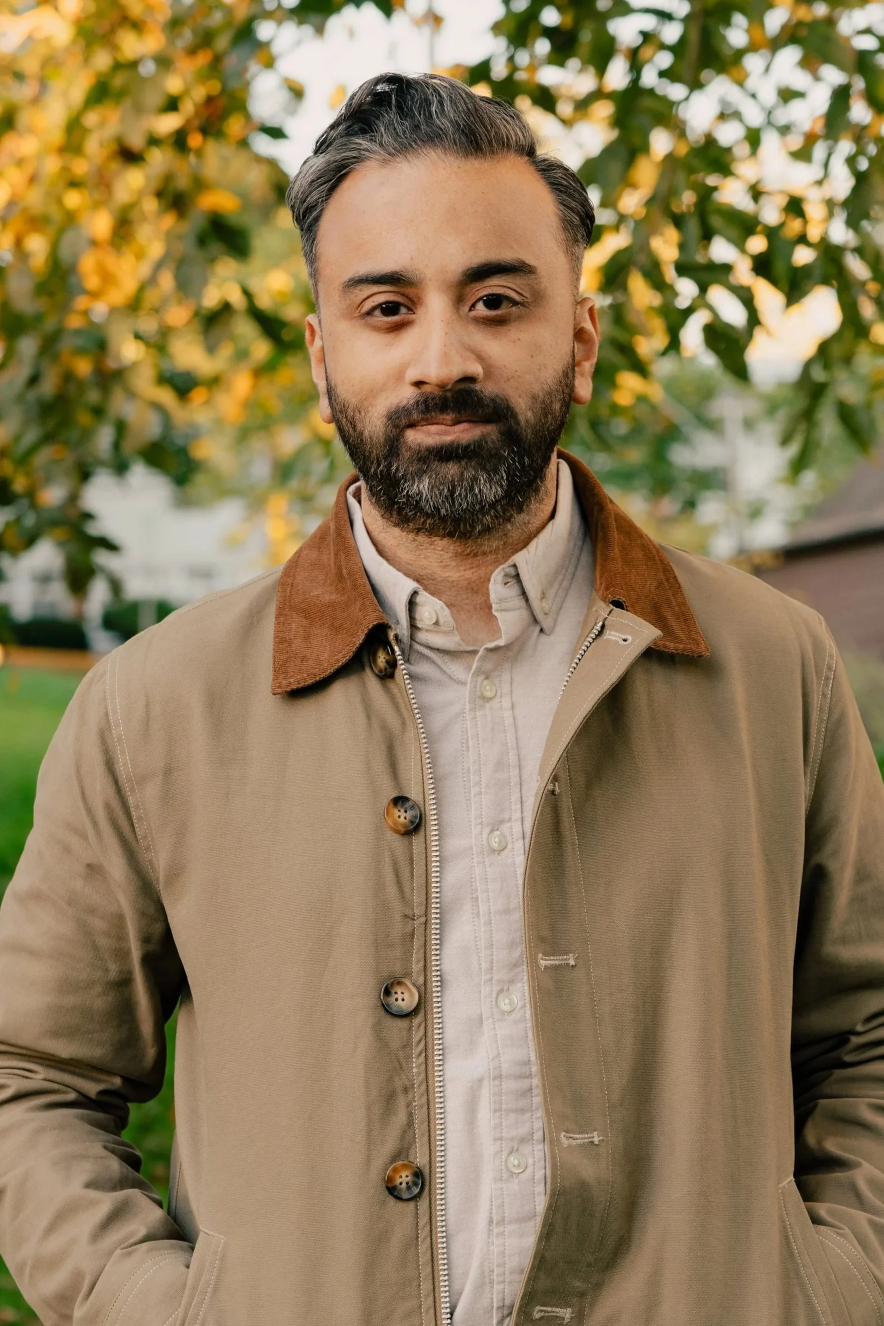 Bearded man in a tan jacket and button-down shirt posing outdoors against blurred autumn foliage