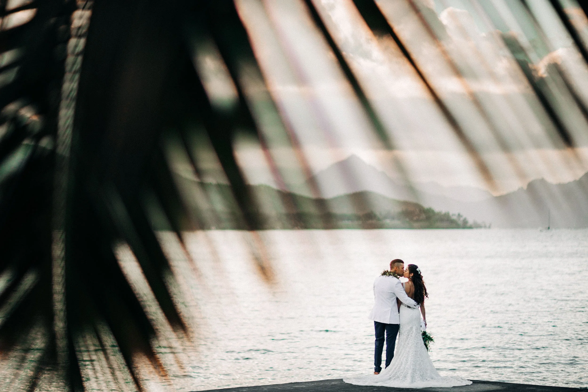 Bride and groom sharing a kiss on a dock in Hawaii at sunset, framed by palm leaves.
