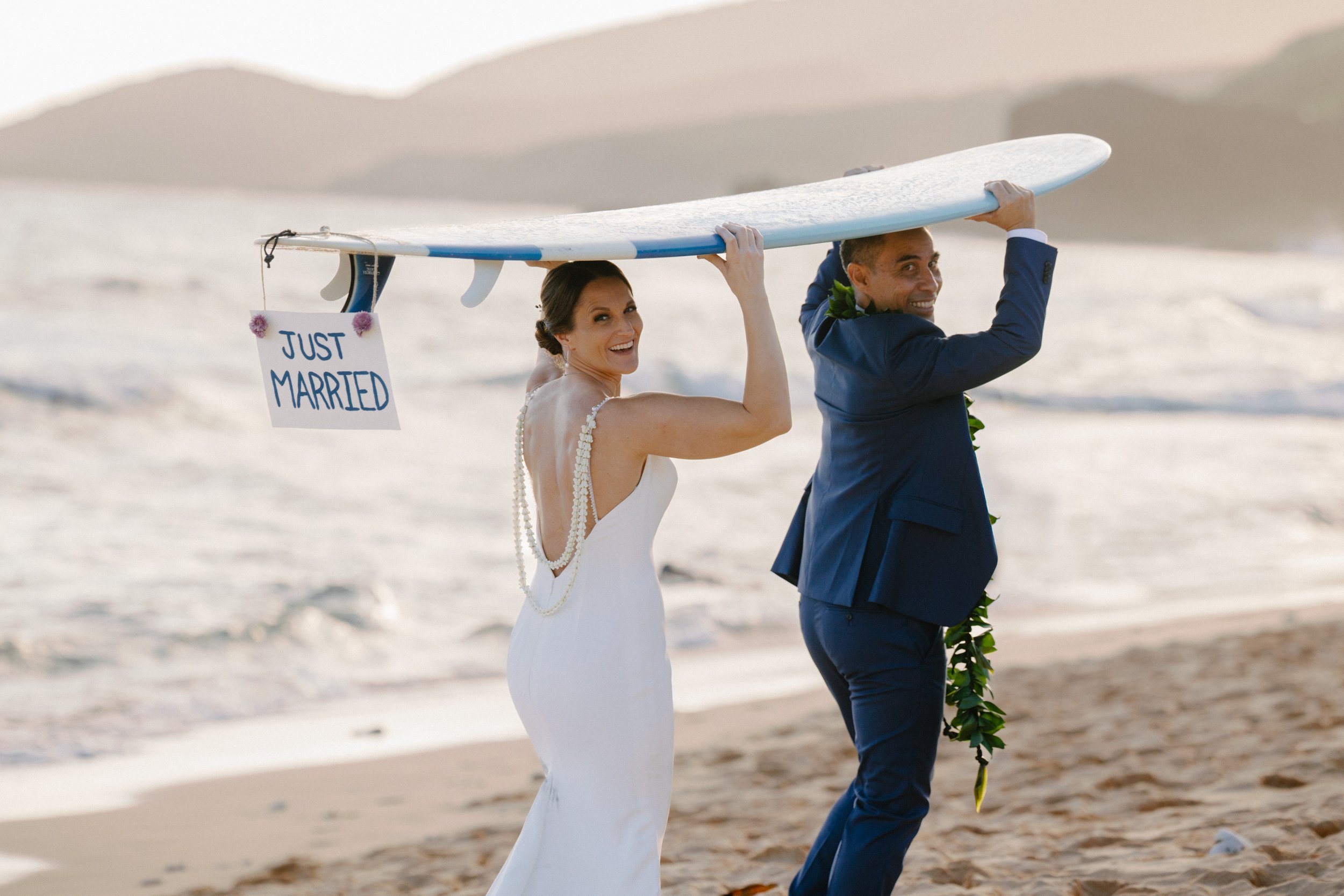 Bride and groom carrying a "Just Married" surfboard on an Oahu beach, captured by an Oahu wedding photographer.