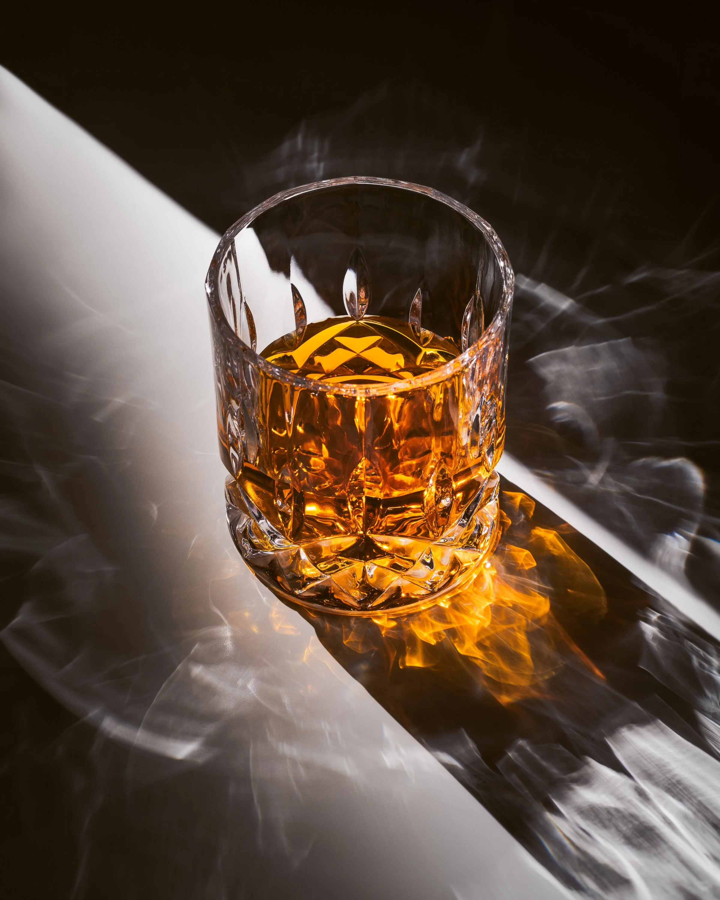 three-quarter view of whiskey in a crystal tumbler being lit from behind by a shaft of light resulting in complex shadows and refraction