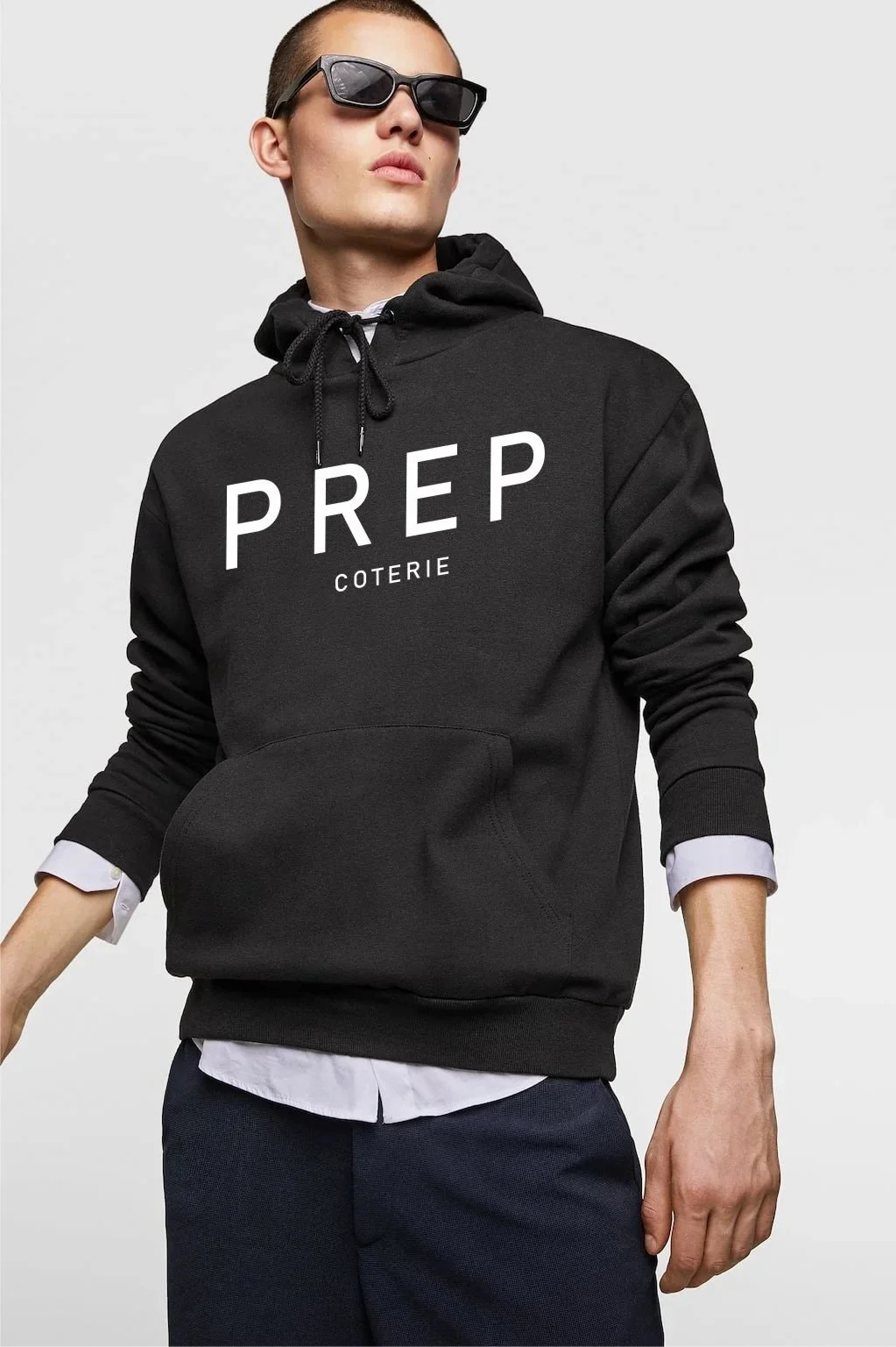 A young man with short hair wearing black sunglasses, a black hoodie with the words "PREP COTERIE" on it, a white shirt underneath, and dark pants.