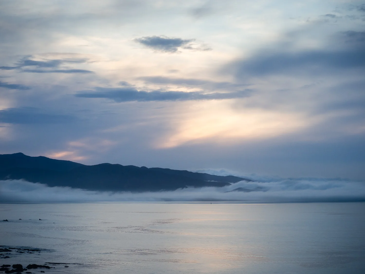 Looking toward the headland extending from the Orongorongo range, on Wellington's south coast. The light of sunrise is reflected in the calm water, but the lighthouse beyond is obscured by the low lying cloud.