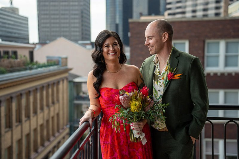 0013_A couple enjoying a moment on a balcony with the cityscape in the background The woman holds a bouquet wearing a red dress while the man is in a green suit.jpg