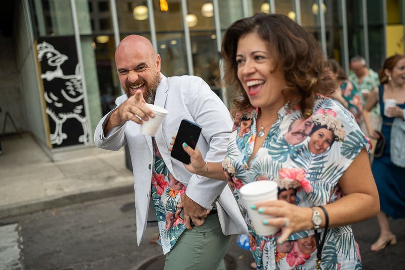 0032_Two people wearing matching tropical shirts and holding drinks share a laugh while walking outside.jpg