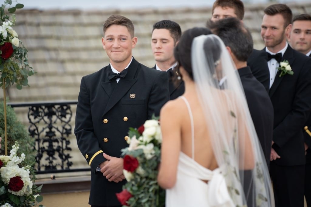 The groom smiles as he sees the bride approaching surrounded by groomsmen in tuxedos and flowers decorating the scene.jpg