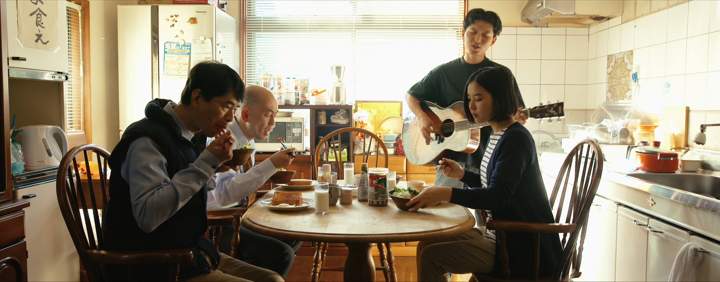 Four people having breakfast in a kitchen. Two men are sitting at a wooden dining table eating with chopsticks, one woman is seated also eating, and a man standing playing a guitar.