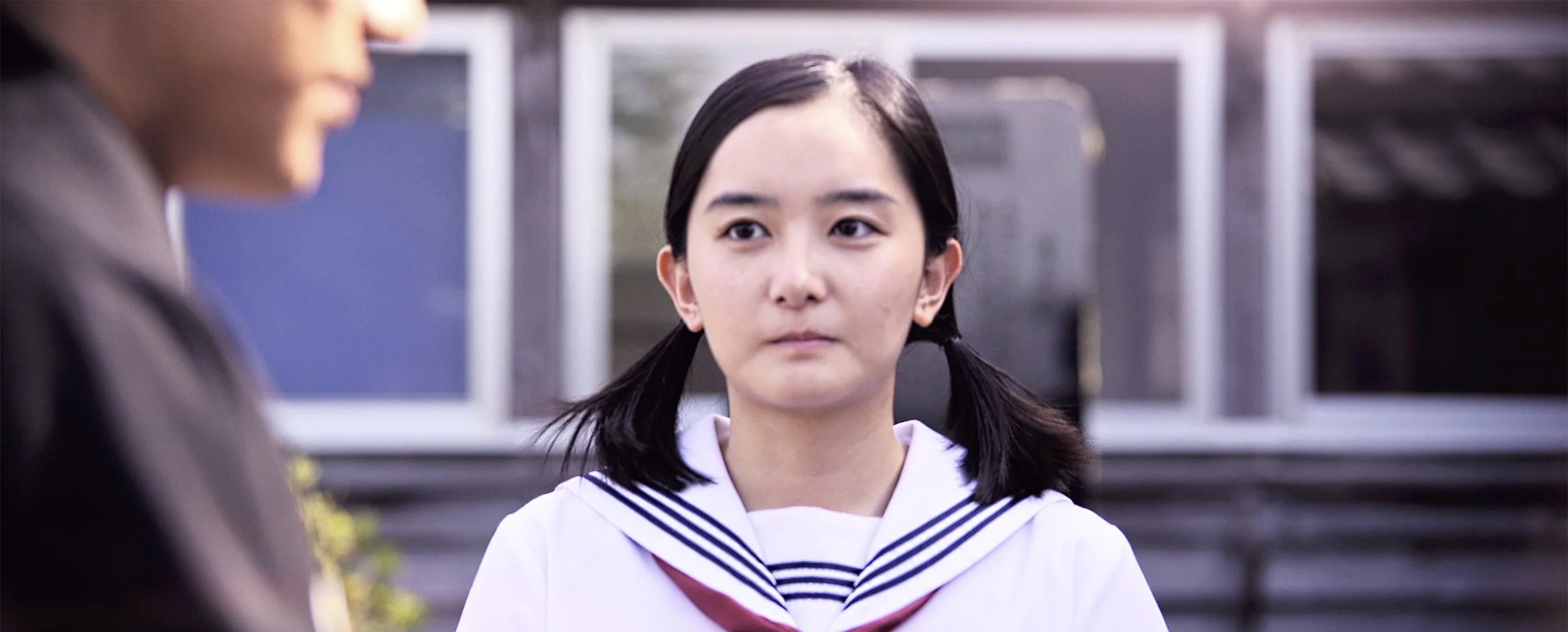 A young woman with black hair in pigtails wearing a white sailor school uniform looking at another person outside with blurred background of school building and windows.