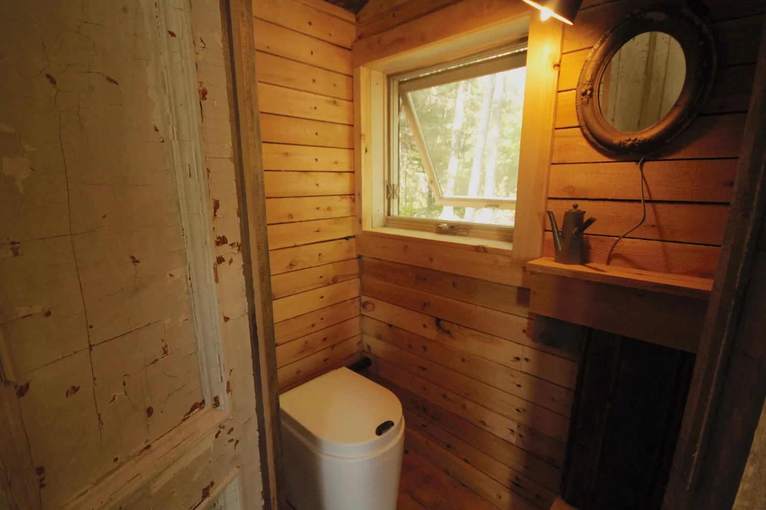 La salle de bain&nbsp;: toilette &eacute;cologique, un miroir rond au mur et une fen&ecirc;tre sur les arbres