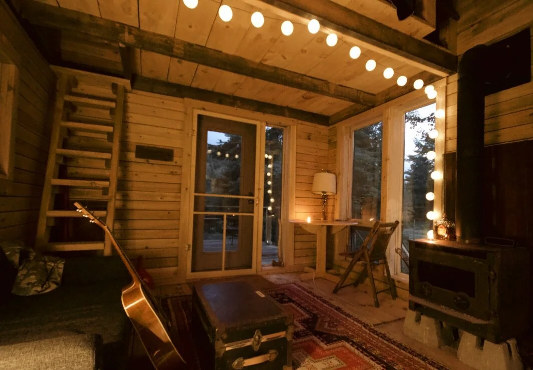 La pi&egrave;ce principale au cr&eacute;puscule, guirlandes allum&eacute;es, po&ecirc;le &agrave; bois qui br&ucirc;le, guitare appuy&eacute;e sur le divan