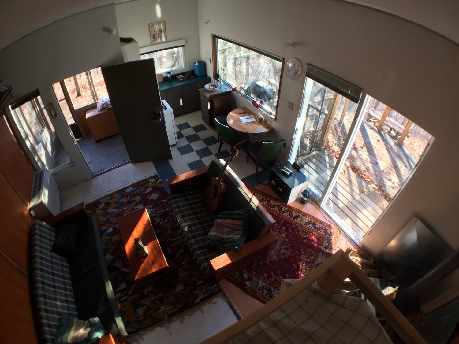 Vue de la mezzanine sur le séjour, tapis, divan, table ronde et cuisine, avec la galerie à travers les fenêtres