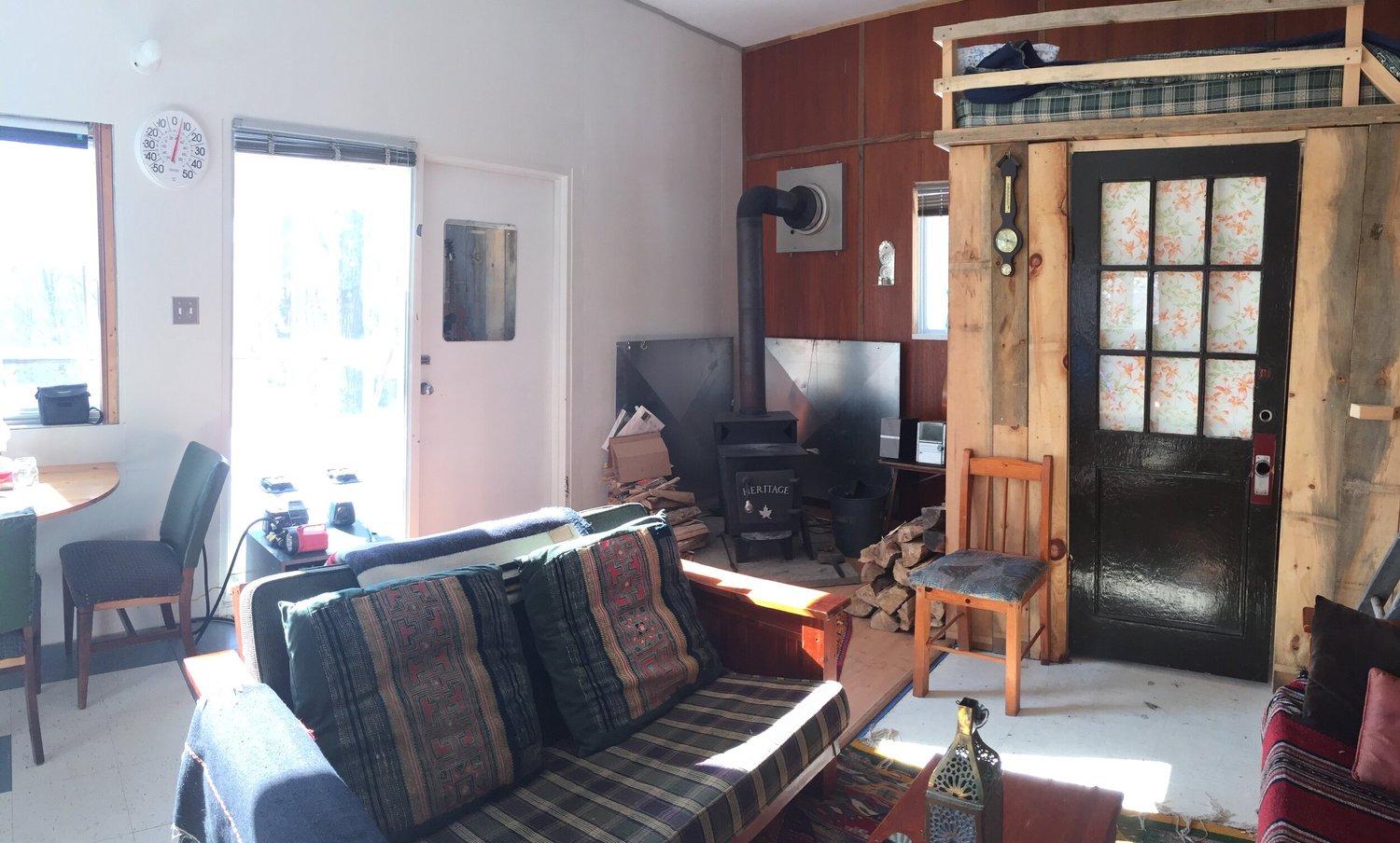 Le séjour : un poêle à bois noir en fonte avec du bois fendu cordé à côté, un divan à l’avant-plan et la mezzanine au-dessus de la porte