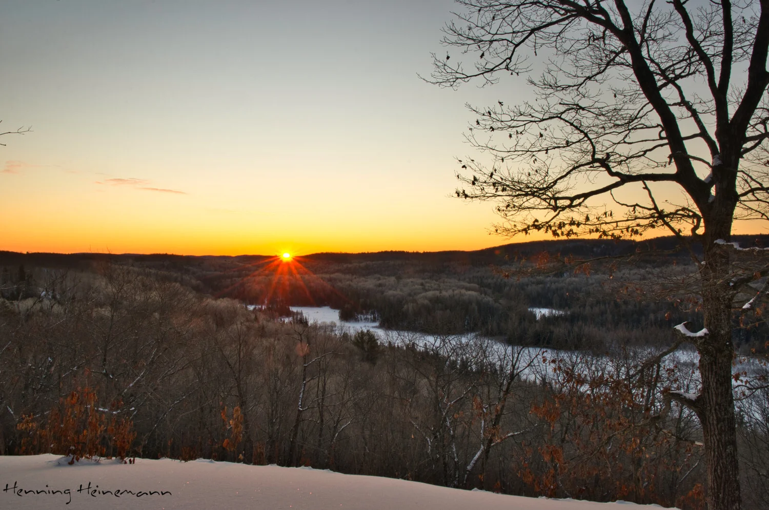 Lever de soleil d’hiver depuis la falaise : le soleil en étoile sur les collines lointaines, vallée enneigée et arbres nus en bas