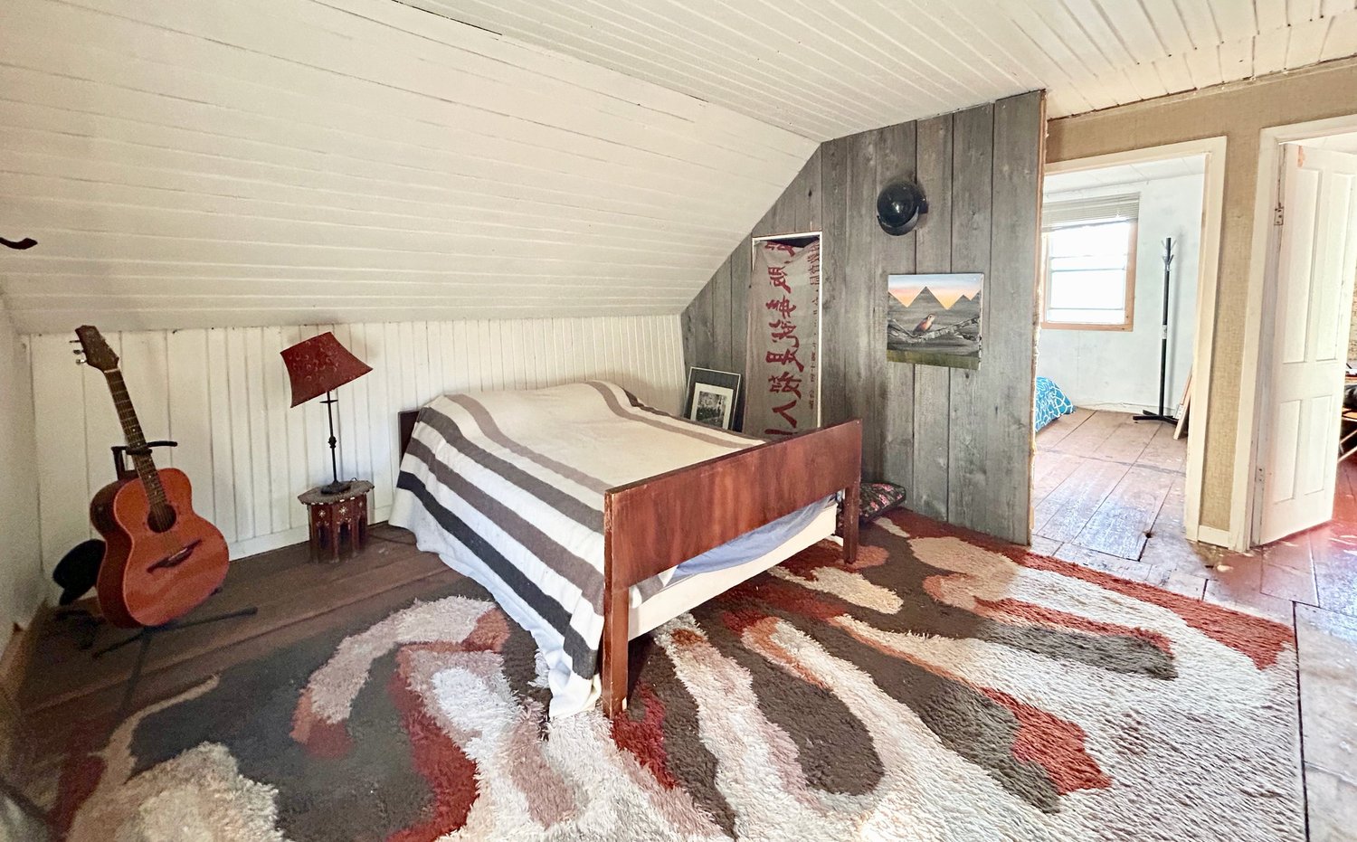 Chambre avec un lit contre un mur en bois de grange sous un plafond en pente, tapis sur un plancher de larges planches, guitare dans le coin